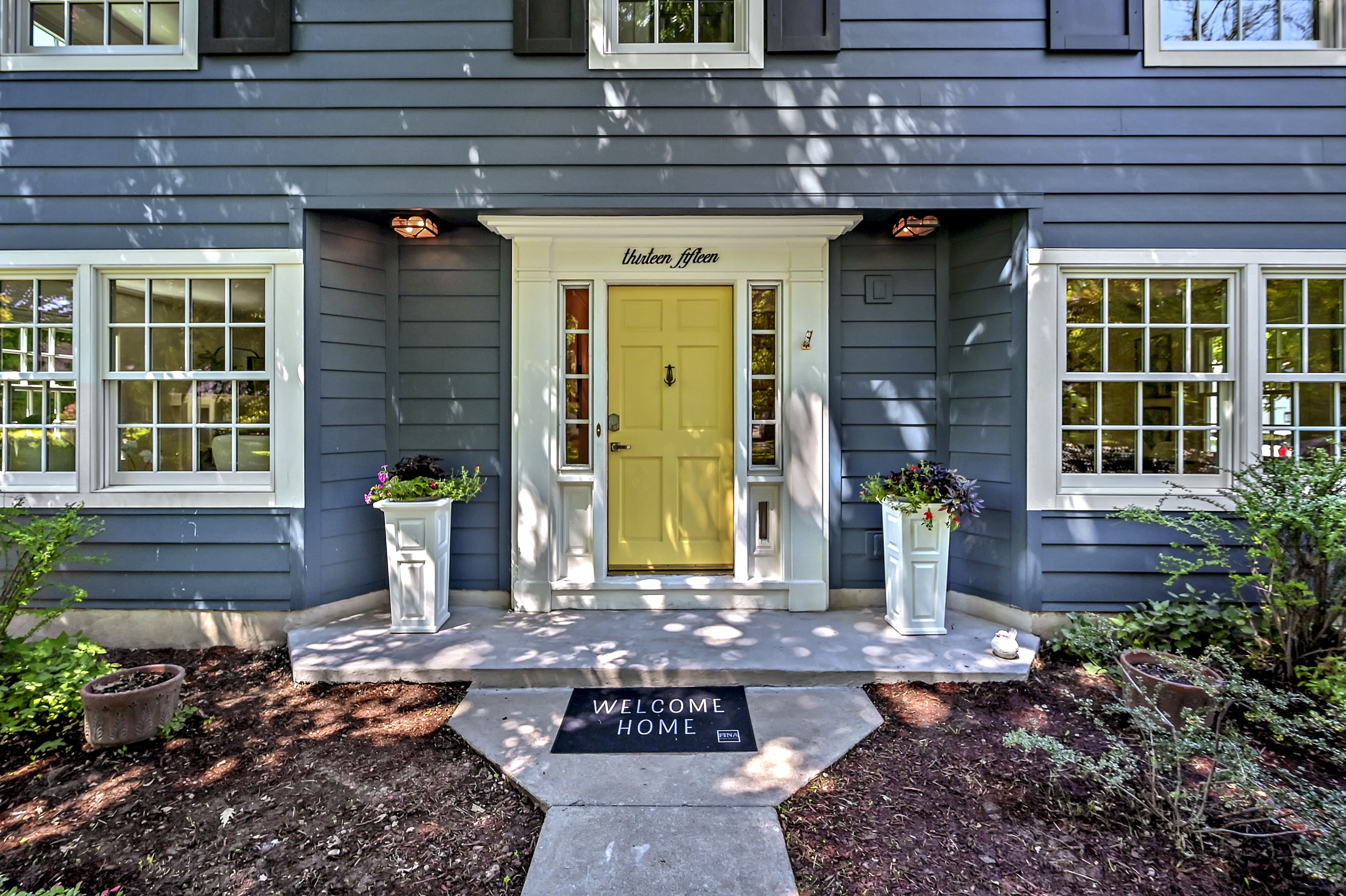 Harrison Front Door Yellow front door on grey house with bright potted flowers and mat that says 'welcome home'