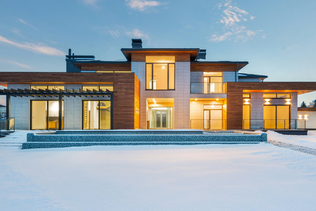 Modern luxury home with large windows and warm lighting, showcasing contemporary architecture with wooden and stone elements. The property is surrounded by snow, highlighting its sleek design and inviting ambiance.