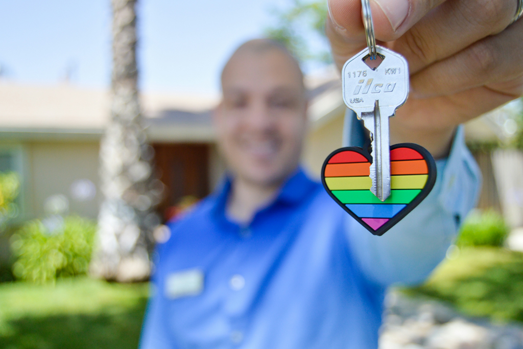 Realtor standing in front of a newly purchased home holding a silver house key with a rainbow heart keychain