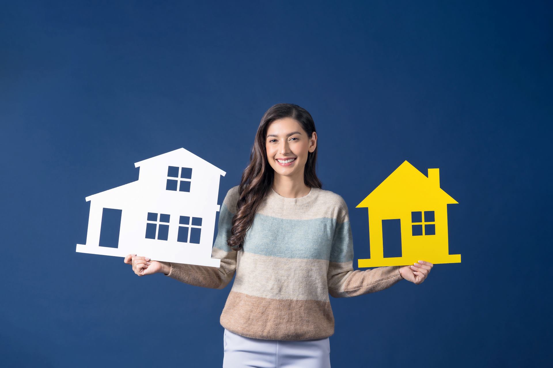 An Asian woman smiles while holding small paper houses in her hands, showcasing creativity and joy.