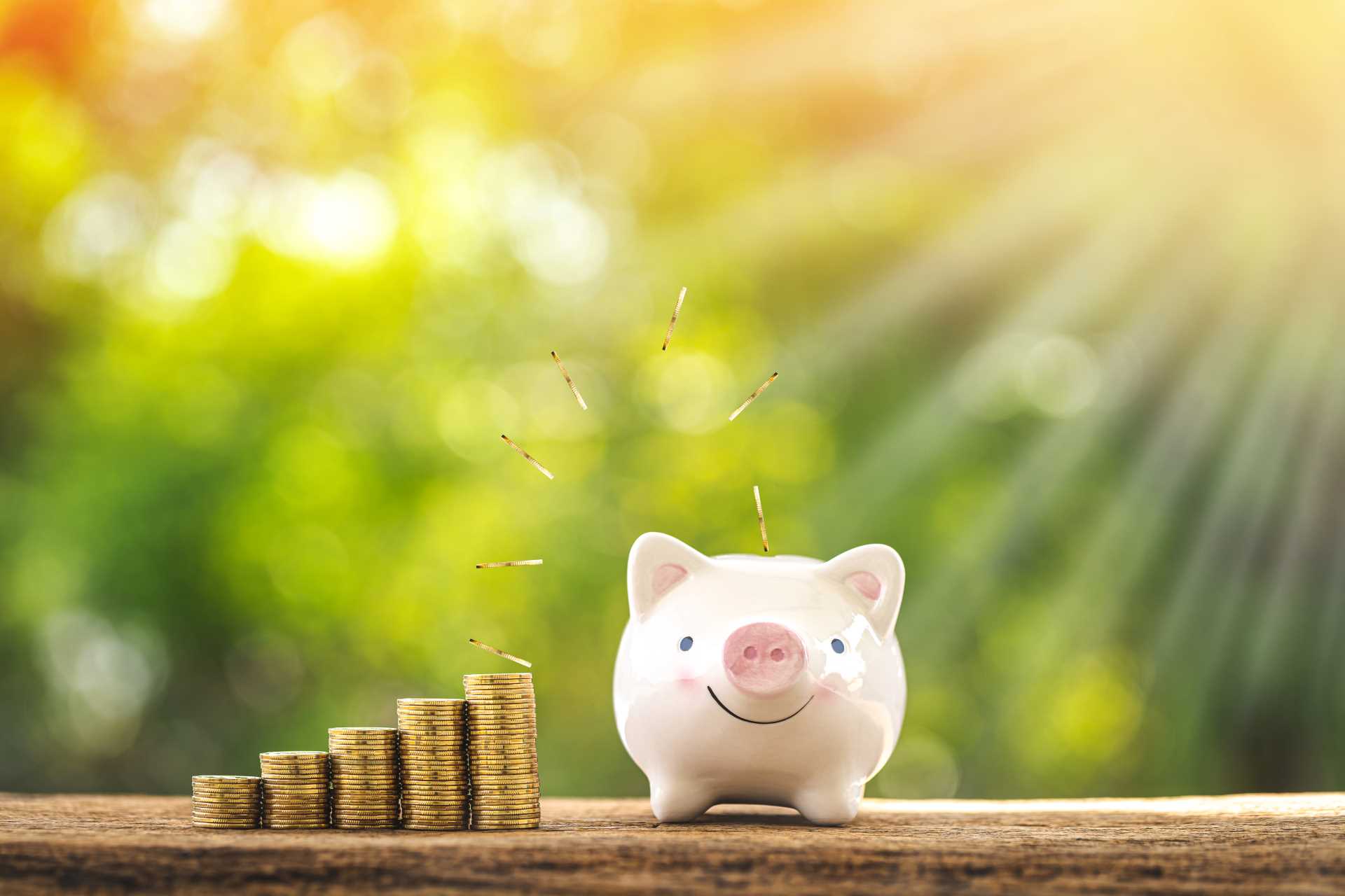 A piggy bank surrounded by coins on a wooden table, illuminated by warm sunlight streaming in.