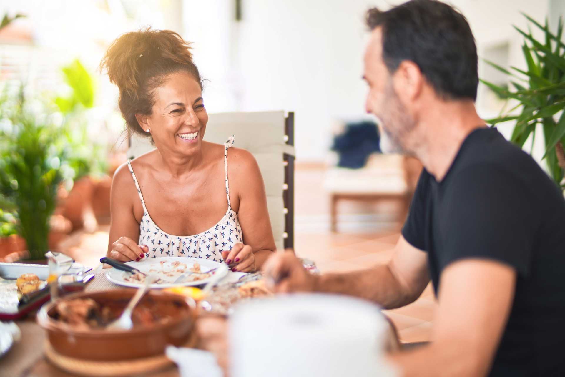 A woman and a man are seated at a table outdoors, smiling at each other. Plates with food are in front of them. Lush, green plants are visible in the background.