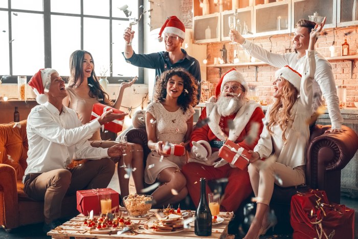 Hosting holiday gatherings in a cozy loft apartment with exposed brick walls, featuring a group of friends celebrating Christmas together on a leather sofa, exchanging wrapped gifts, wearing Santa hats and festive attire, with champagne glasses raised, confetti falling, and a coffee table spread with snacks, drinks, and candles creating a warm intimate party atmosphere.