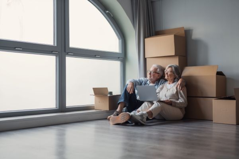 Downsizing in Dallas. Senior couple sitting on the floor beside moving boxes in a bright modern home, using a laptop while planning their retirement move and downsizing.