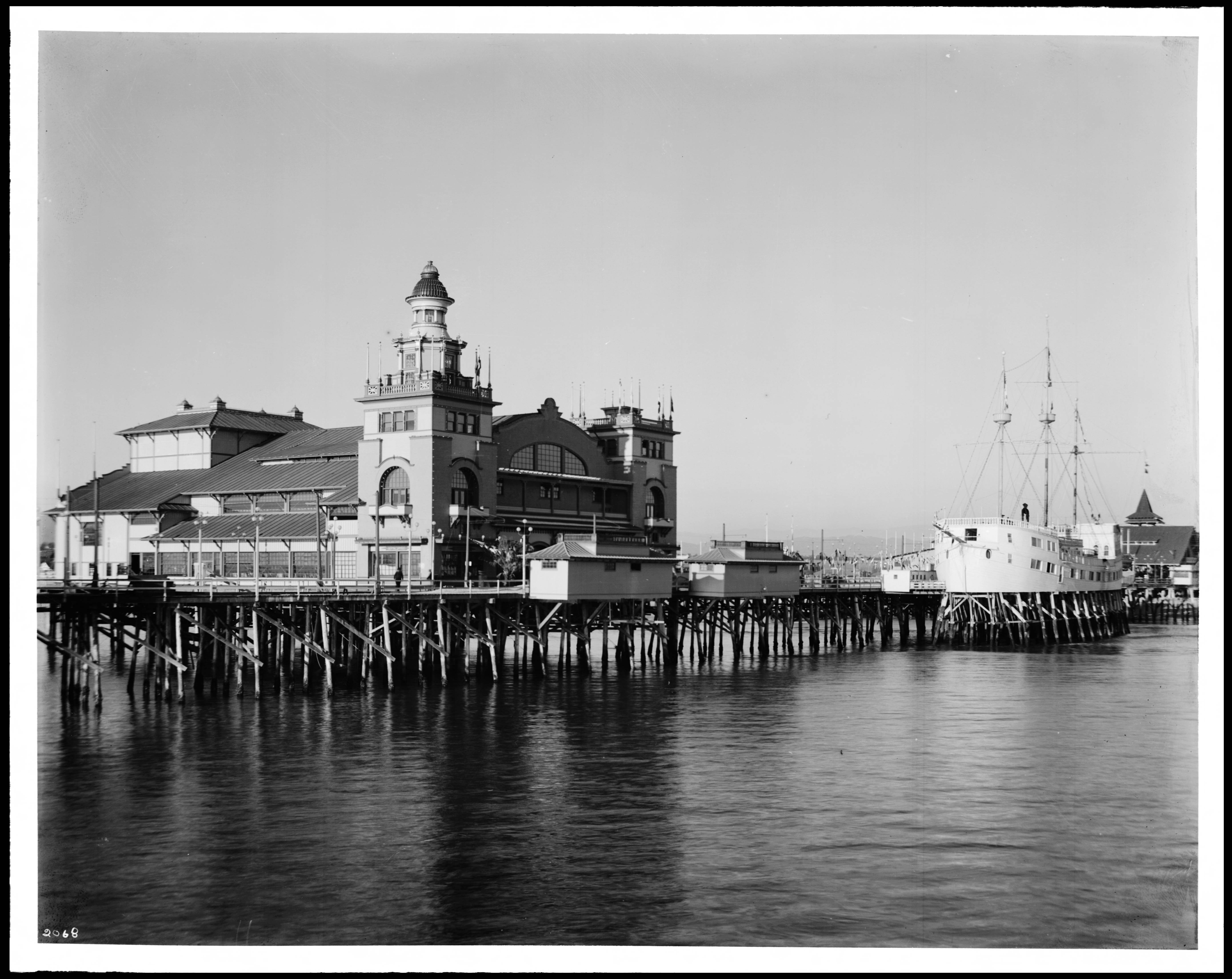 Black-and-white historic photograph of the Venice wharf and pavilion in 1913, with large buildings on pilings over the water and a ship docked beside the pier.