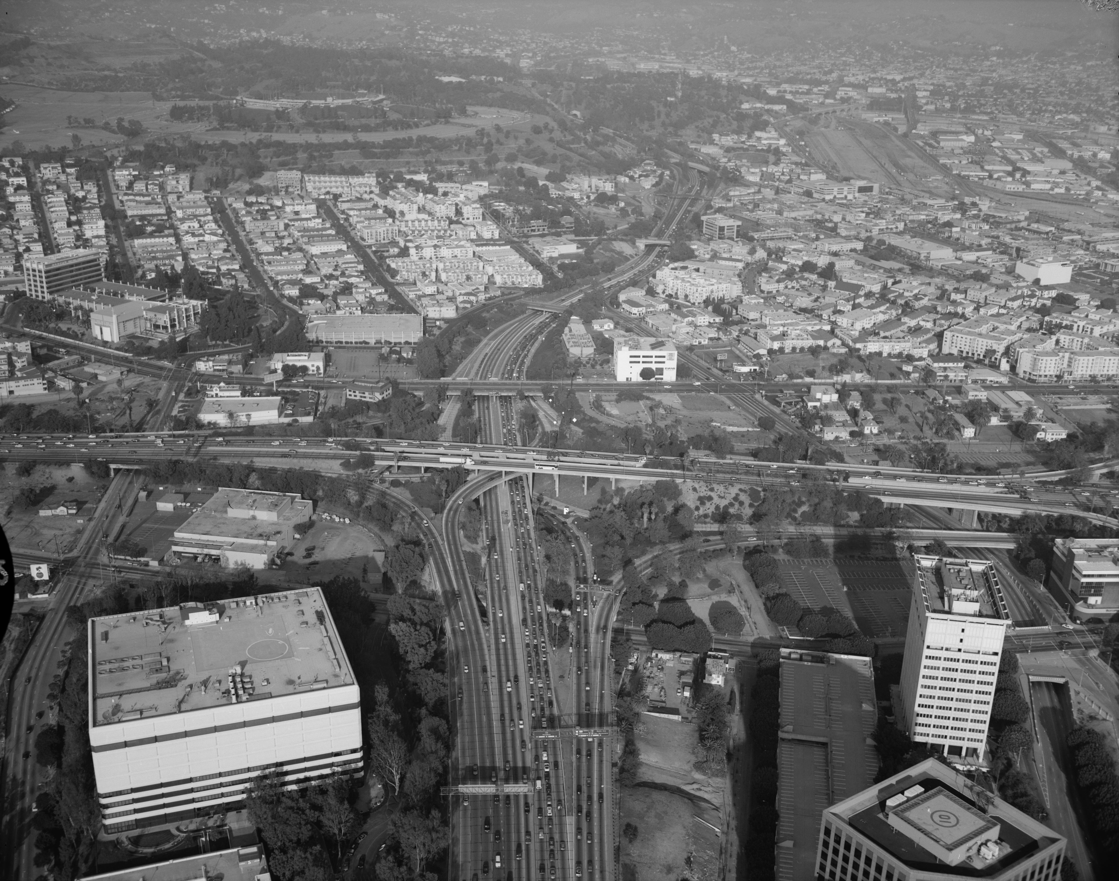 The Four Level Interchange, one of the freeway structures that reshaped movement around Downtown Los Angeles. Aerial black-and-white photograph of the Four Level Interchange near Downtown Los Angeles with multiple freeways, office buildings, and surrounding neighborhoods visible.