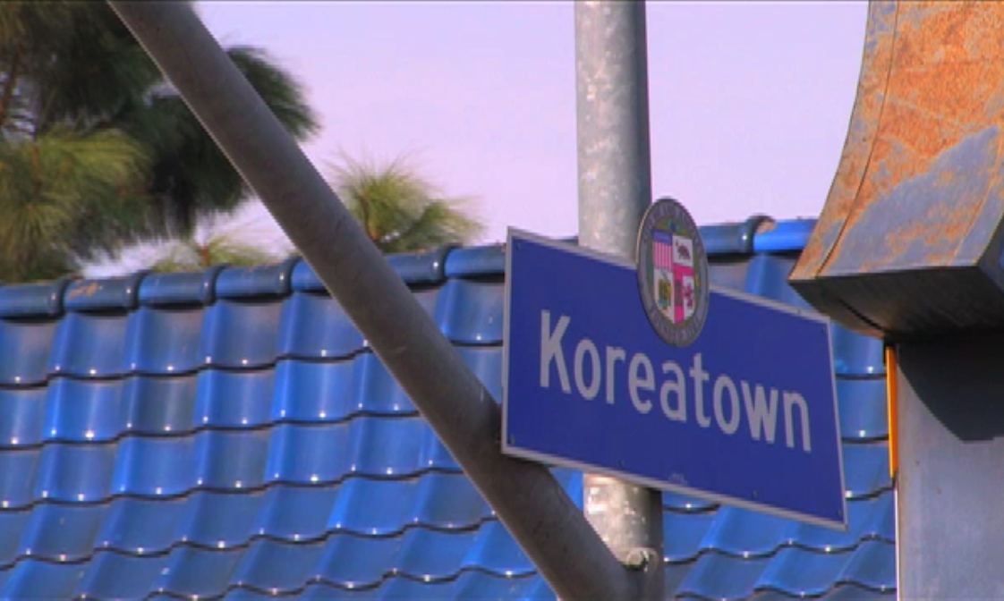 Busy Koreatown street scene with cars, Korean business signs, storefronts and Downtown Los Angeles in the background.