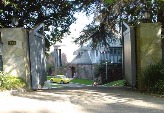 Gated entrance to Zsa Zsa Gabor’s Bel Air home, with stone gateposts, open gates, a driveway, trees and a yellow car near the house.