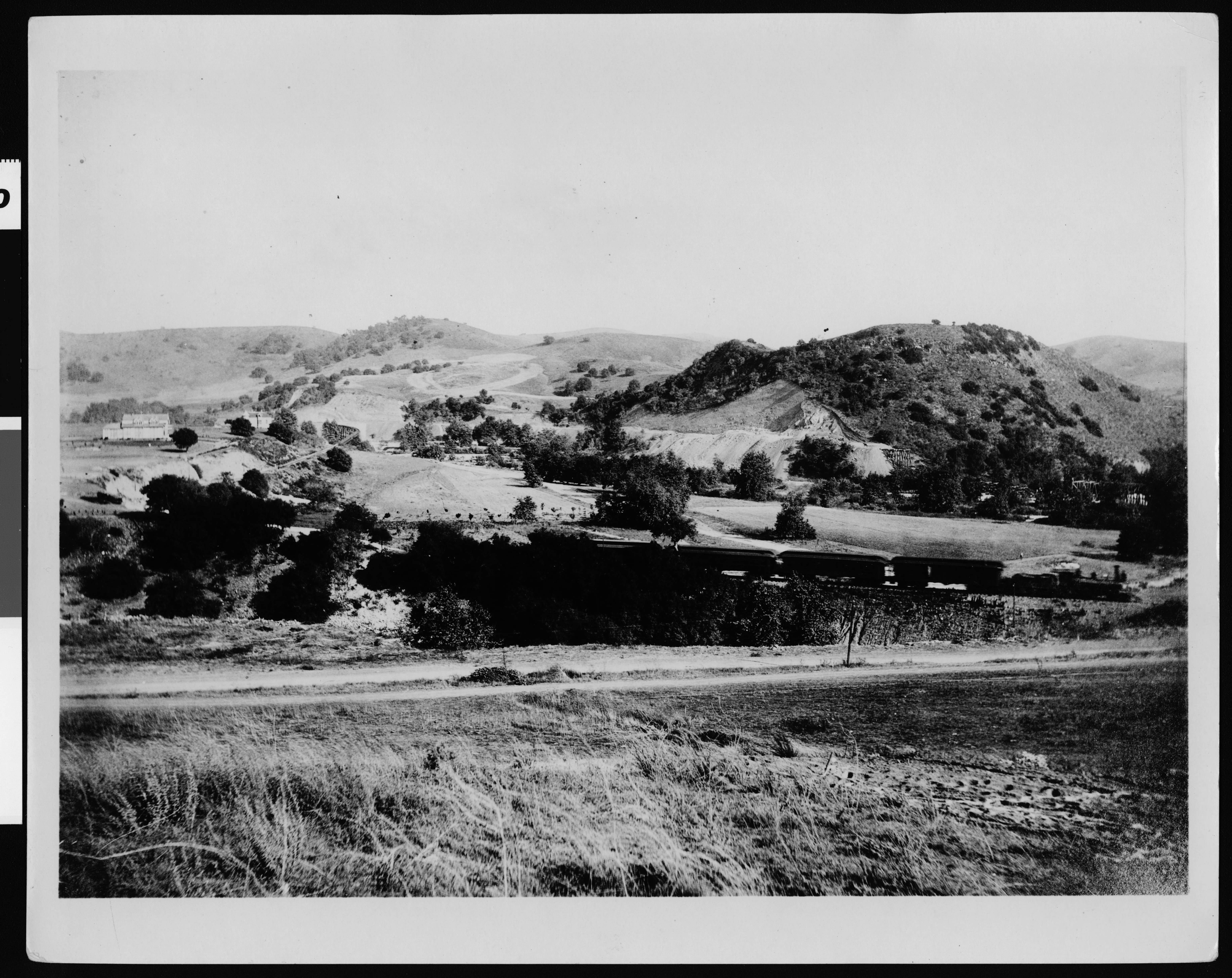 Black-and-white historic photograph of the Arroyo Seco railroad bridge near Highland Park, with open hillsides, tracks and early buildings in the distance.