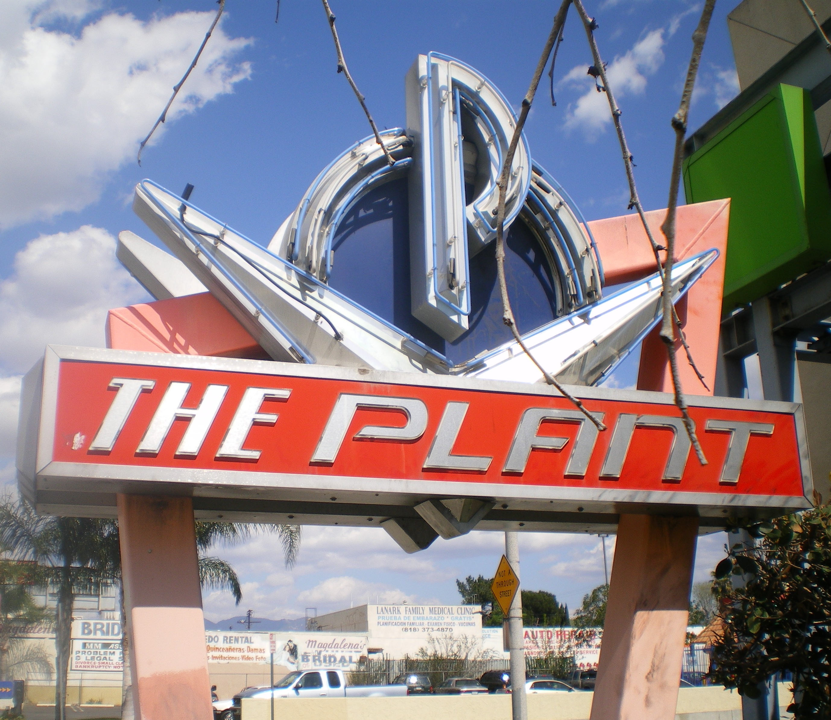 Color photograph of The Plant sign in Van Nuys, with a retro-style neon sign, blue sky, commercial buildings and cars in the background.