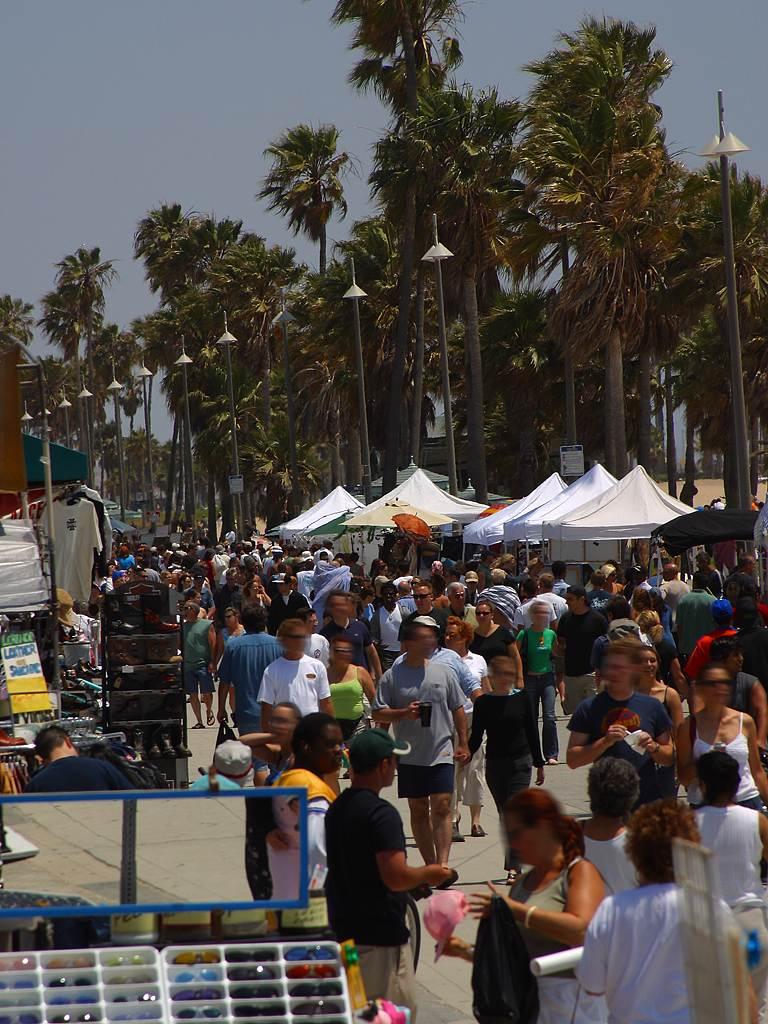 Busy Venice Beach Boardwalk with crowds walking past vendor tents, palm trees, and beachside shops.