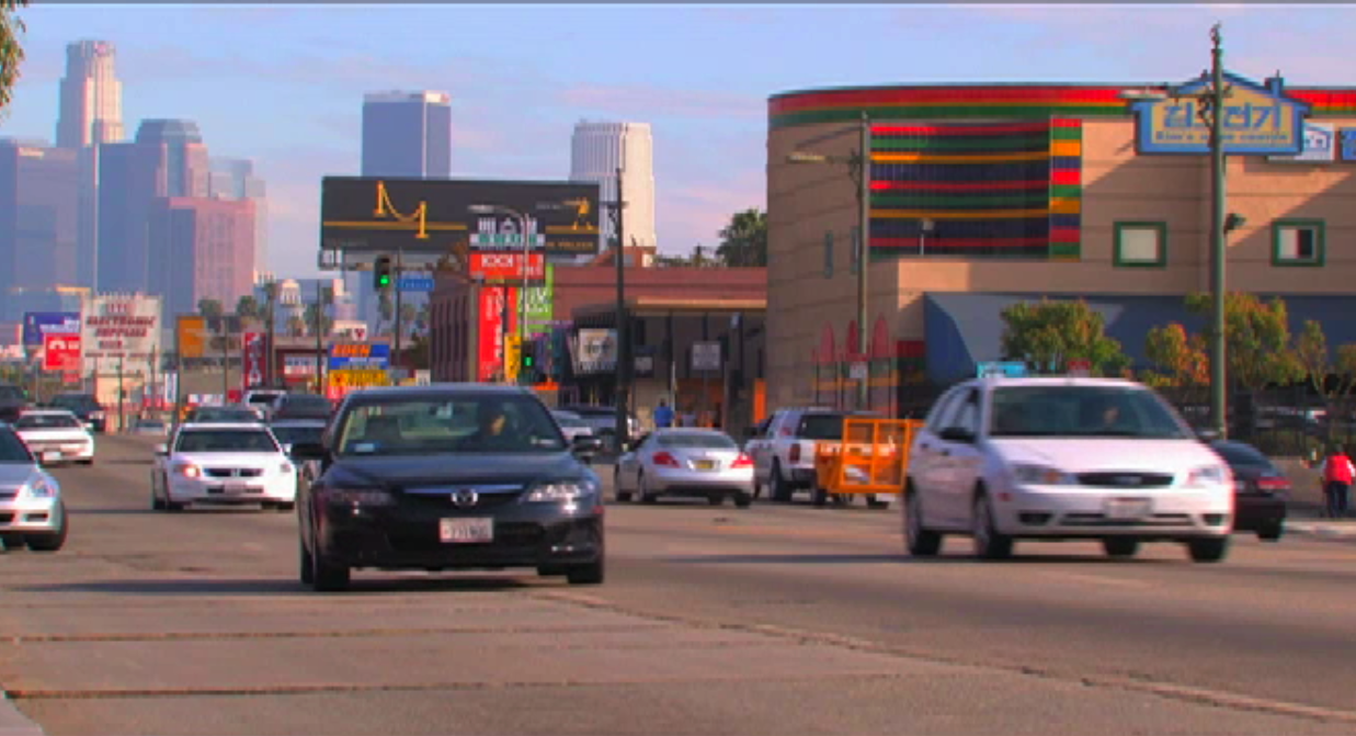 Street view of Olympic Boulevard in Koreatown with Korean businesses, signs, cars and Downtown Los Angeles in the distance.