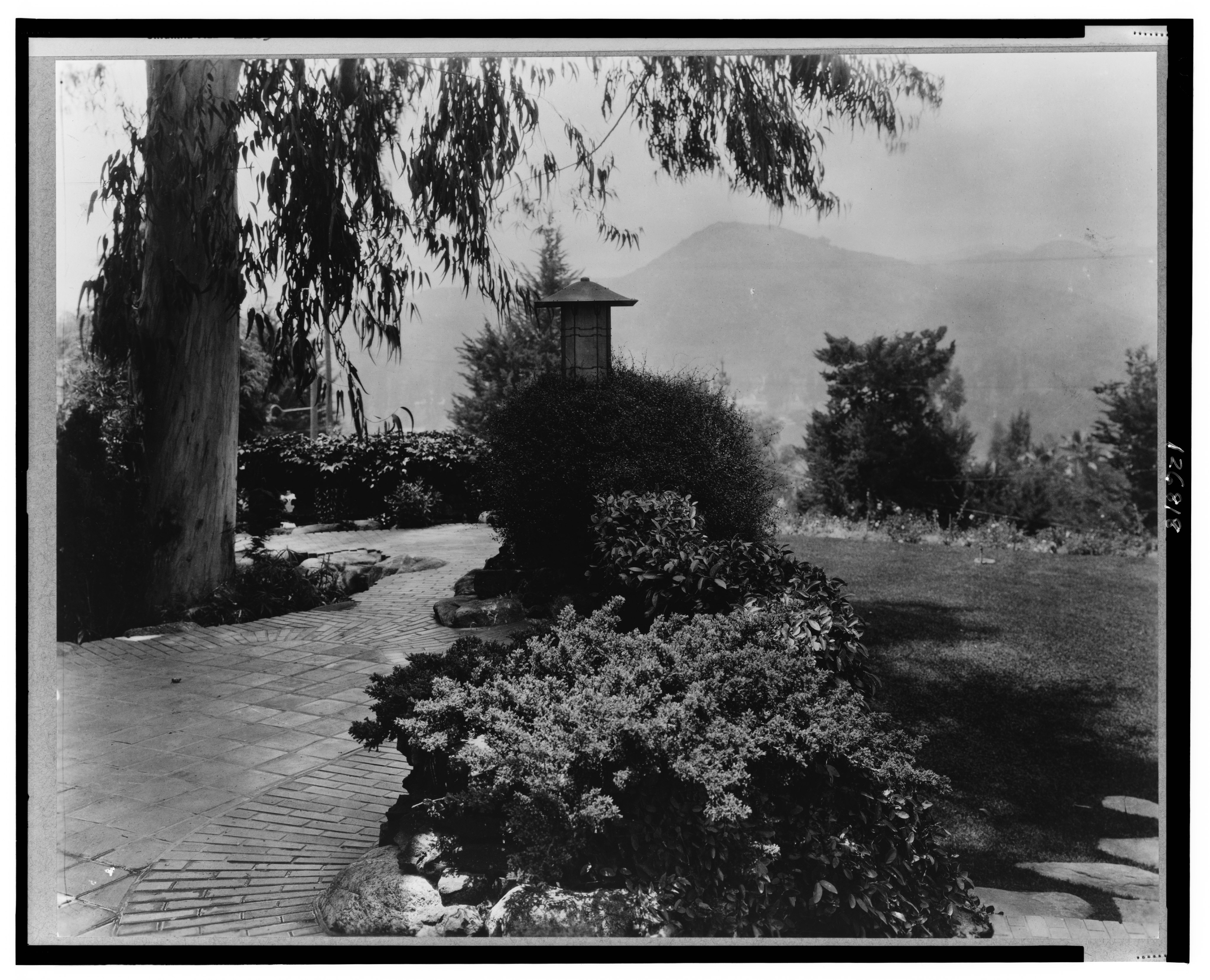 Black-and-white photograph of the Gamble House water terrace in Pasadena, with garden paths, shrubs, trees and mountain views in the background.