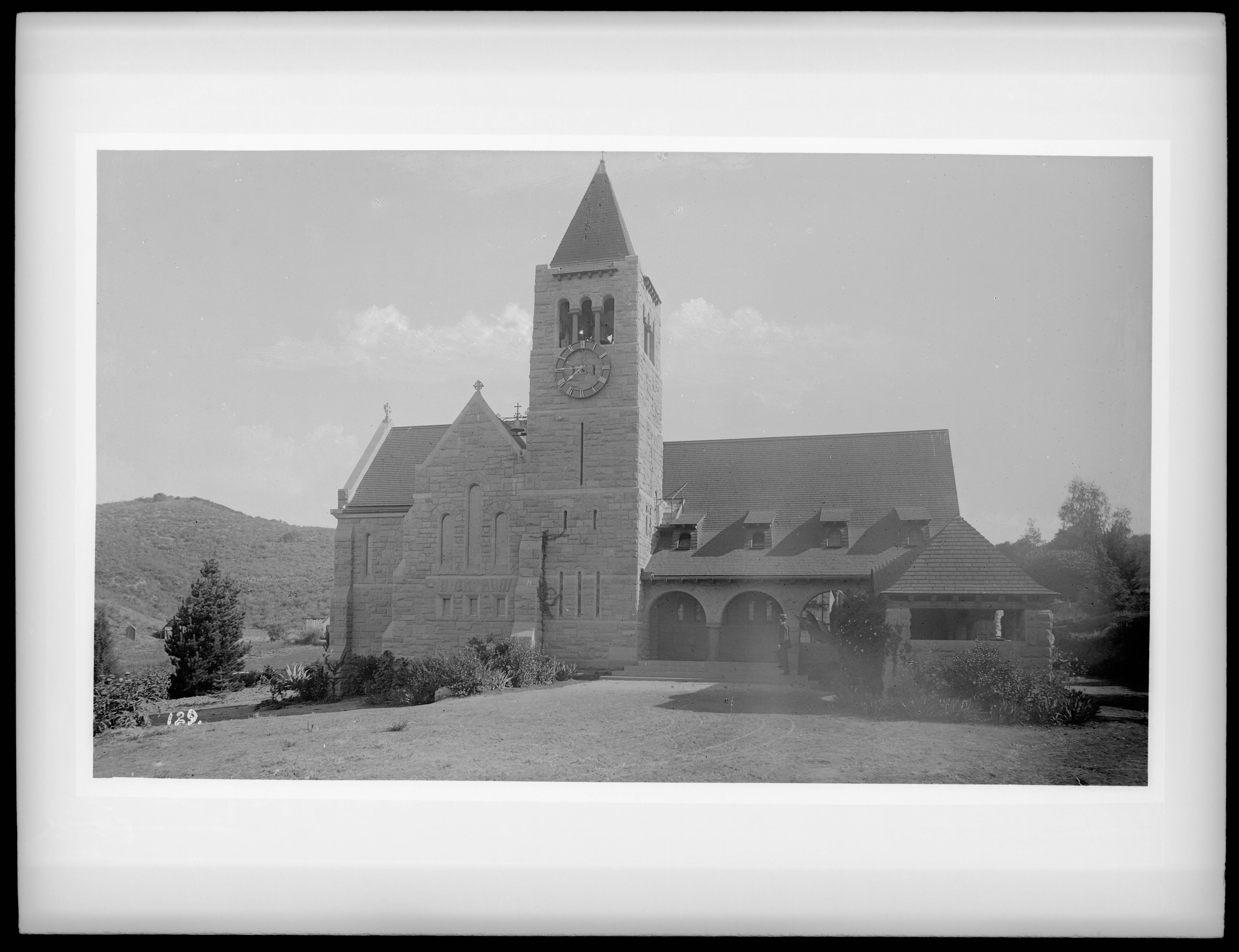 Black-and-white historic photograph of Church of the Angels near Highland Park, with a stone clock tower, arched entry and hills behind it.