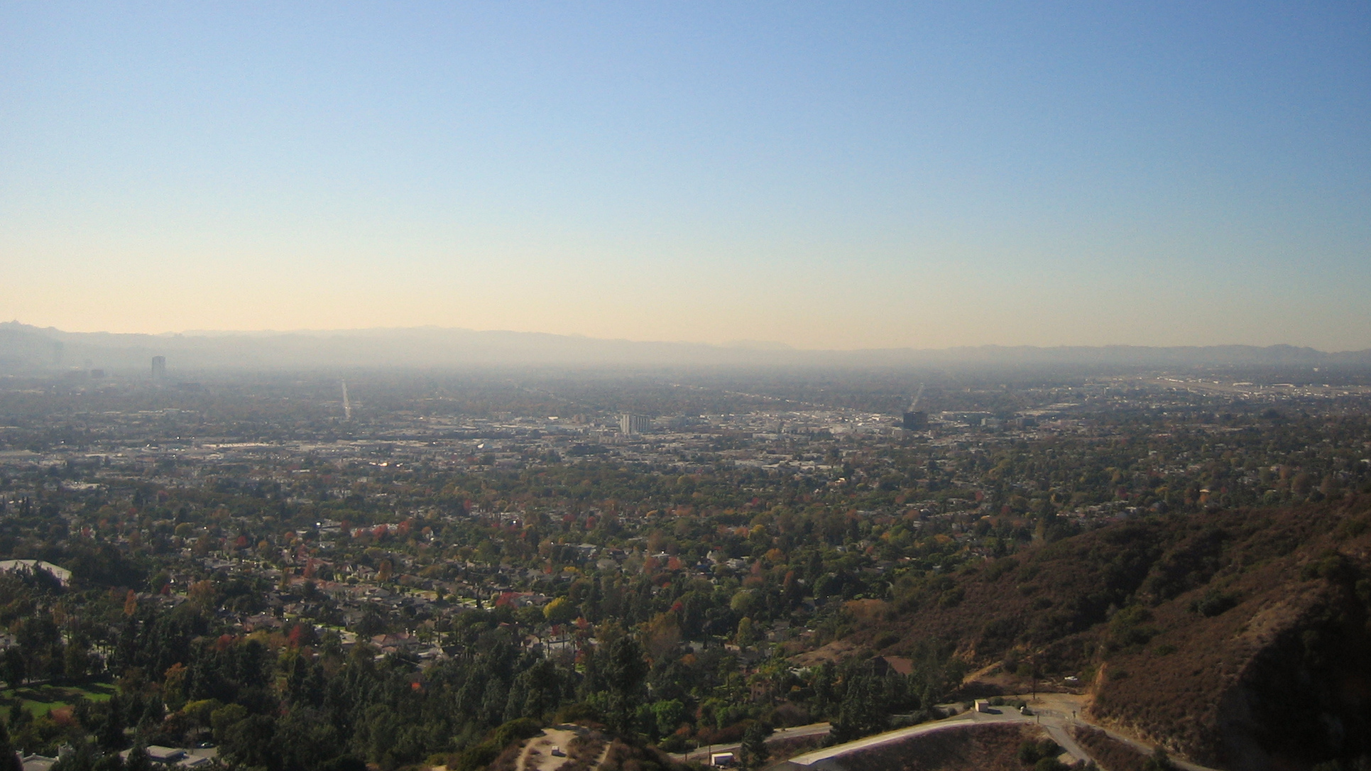 Wide view over the San Fernando Valley from the hills, with neighborhoods, streets, commercial buildings and distant mountains under a hazy sky.