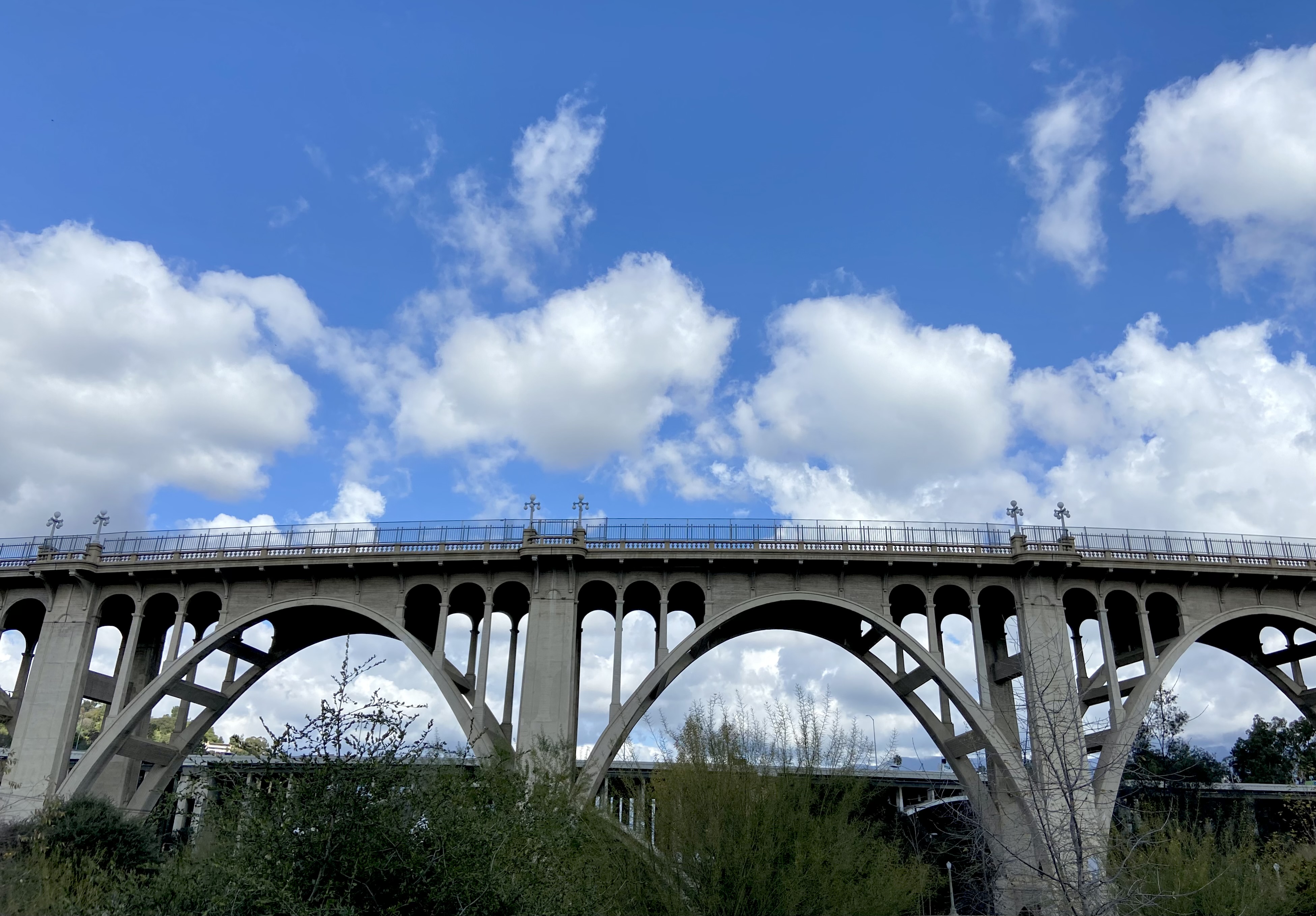Colorado Street Bridge in Pasadena with concrete arches, railings, green vegetation and a blue sky with clouds.