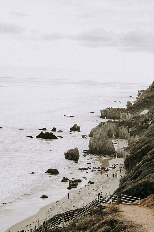 Malibu’s beaches create enormous demand, but coastal land comes with regulations, site constraints, and high development costs. El Matador State Beach in Malibu with rock formations, beachgoers, cliffs, and the Pacific Ocean.