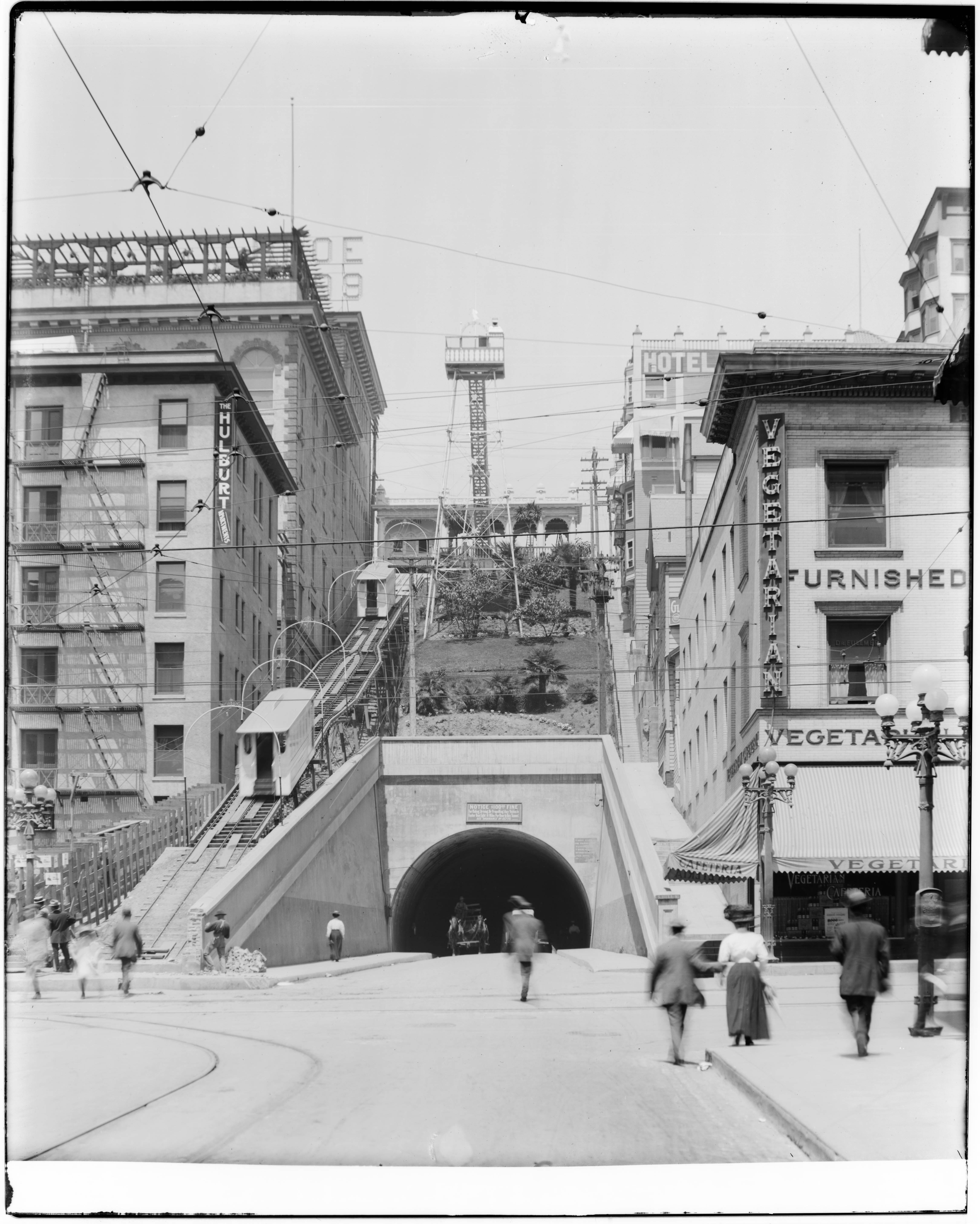 Angels Flight and the Hill Street Tunnel, showing how early Downtown Los Angeles moved through its steep terrain. Black-and-white historic photograph of Angels Flight railway above the Hill Street Tunnel in Downtown Los Angeles with pedestrians, streetcars, hotel signs, and early twentieth-century buildings.