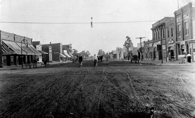 Historic commercial street in Inglewood, showing the city’s early downtown character. Black-and-white historic photograph of a commercial street in early Inglewood, with storefronts, pedestrians, a horse-drawn vehicle, and an unpaved road.