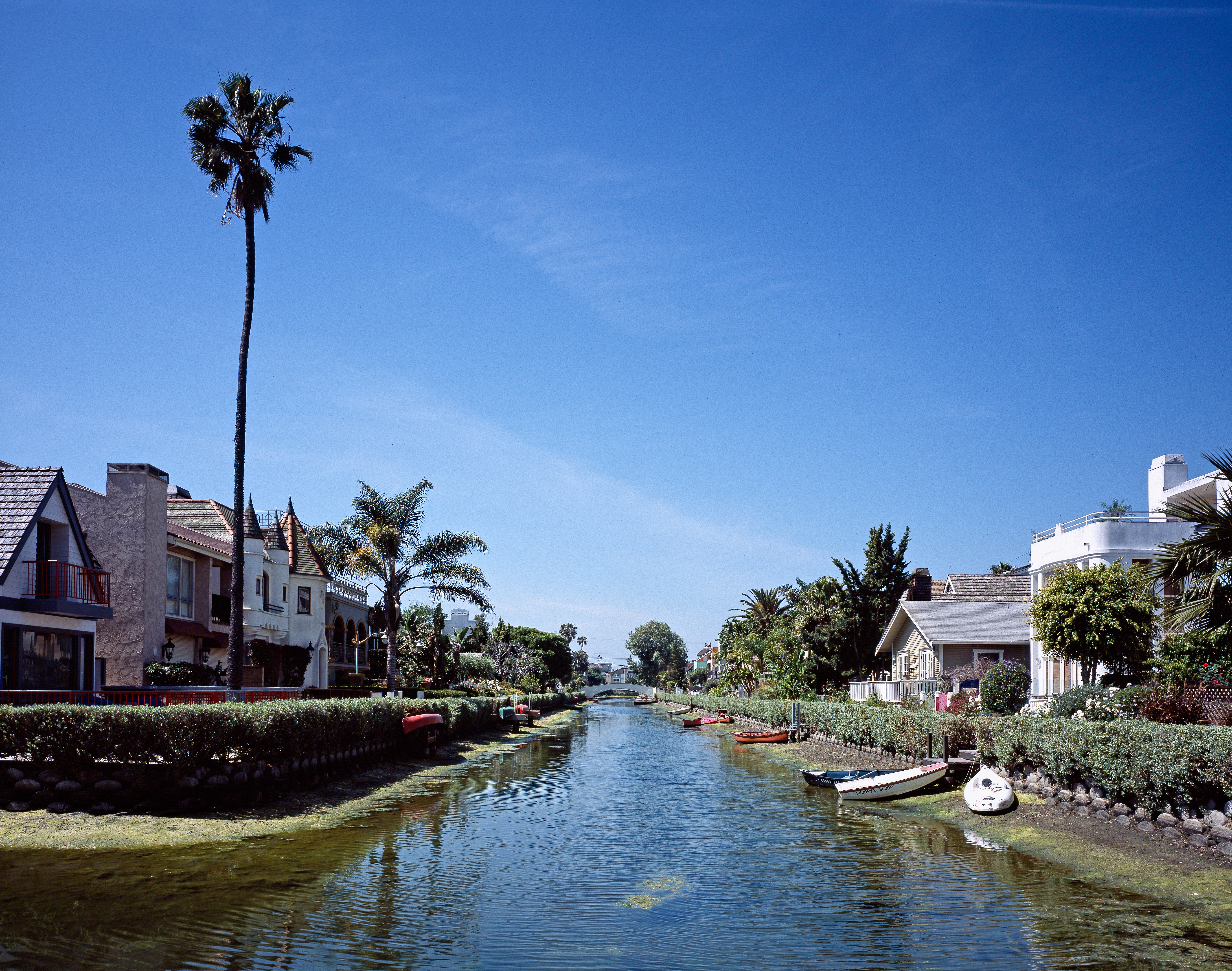 Venice Canals in Los Angeles with waterfront homes, small boats, palm trees, and calm water under a bright blue sky.