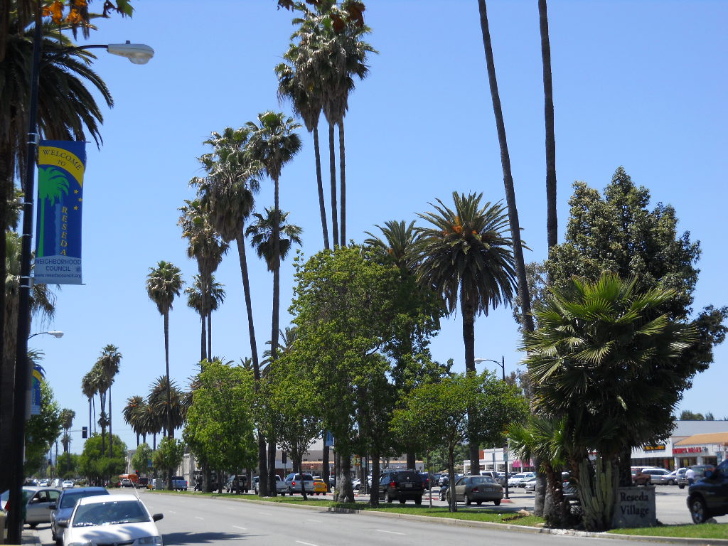 Street view of Sherman Way in Reseda with low-rise storefronts, signs for Latino businesses, parked cars and a clear sky.