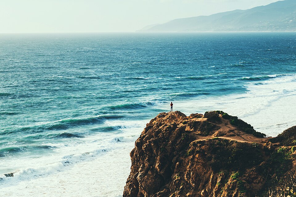Malibu’s coastline is one of the most desirable stretches of real estate in Southern California. Wide ocean view from Point Dume in Malibu, with a person standing on a rocky cliff above the Pacific Ocean.