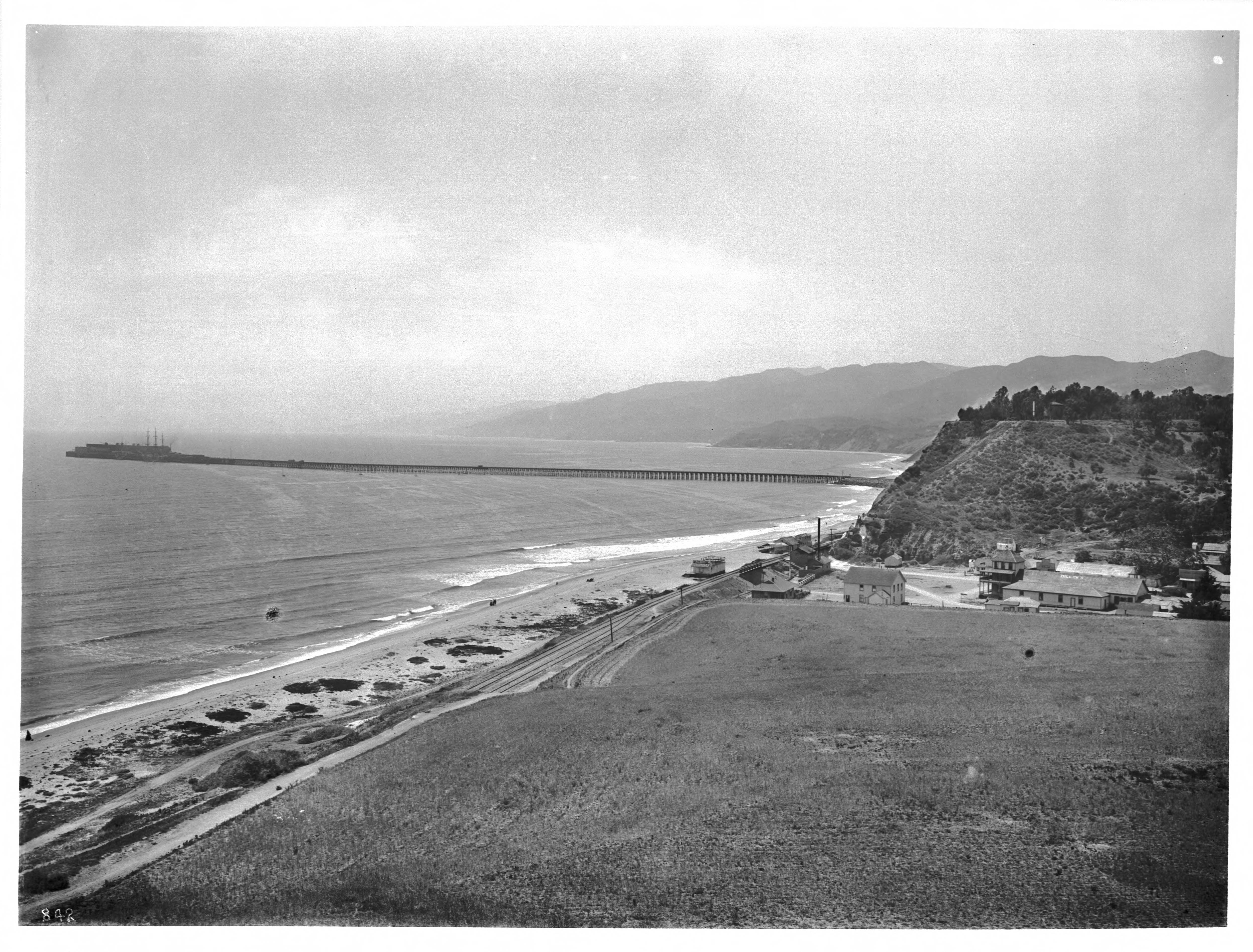 Black-and-white historic photo of Santa Monica Canyon and the Long Wharf of Port Los Angeles around 1900, showing the beach, coastline, railroad tracks, mountains, and a long pier extending into the ocean.