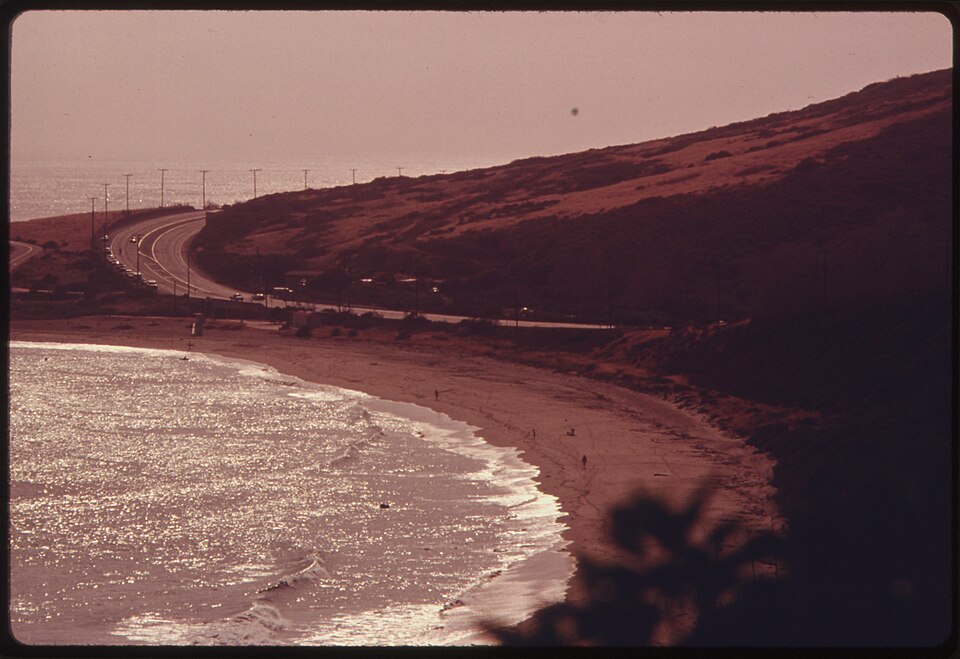 Pacific Coast Highway north of Malibu, a reminder that Malibu’s beauty and access have always been central to its value. Historic color photograph of Pacific Coast Highway running along the Malibu coastline at sunset, with beach, hills, ocean, and cars visible