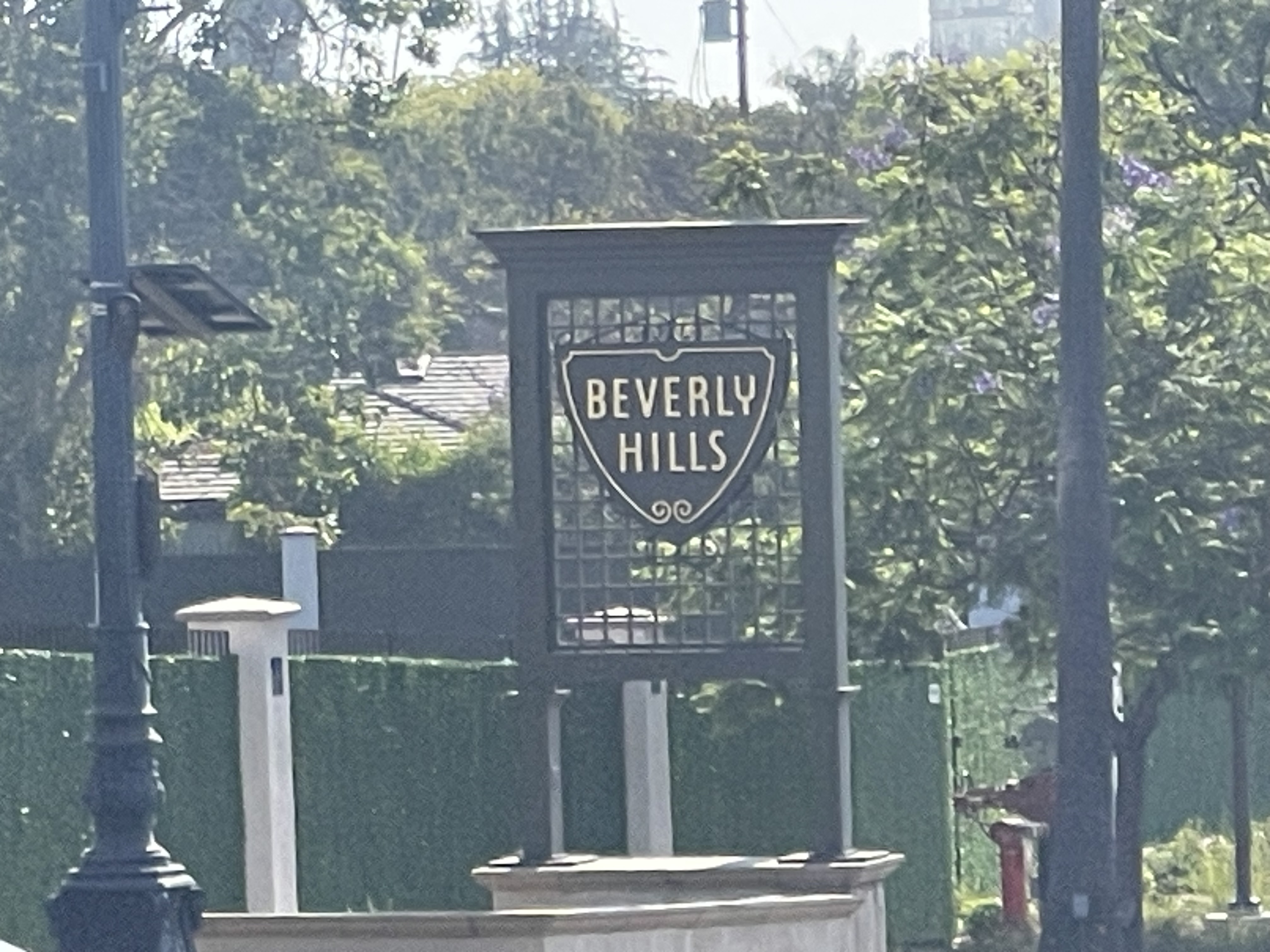 Beverly Hills welcome sign on a landscaped street with trees, hedges and sunlight.