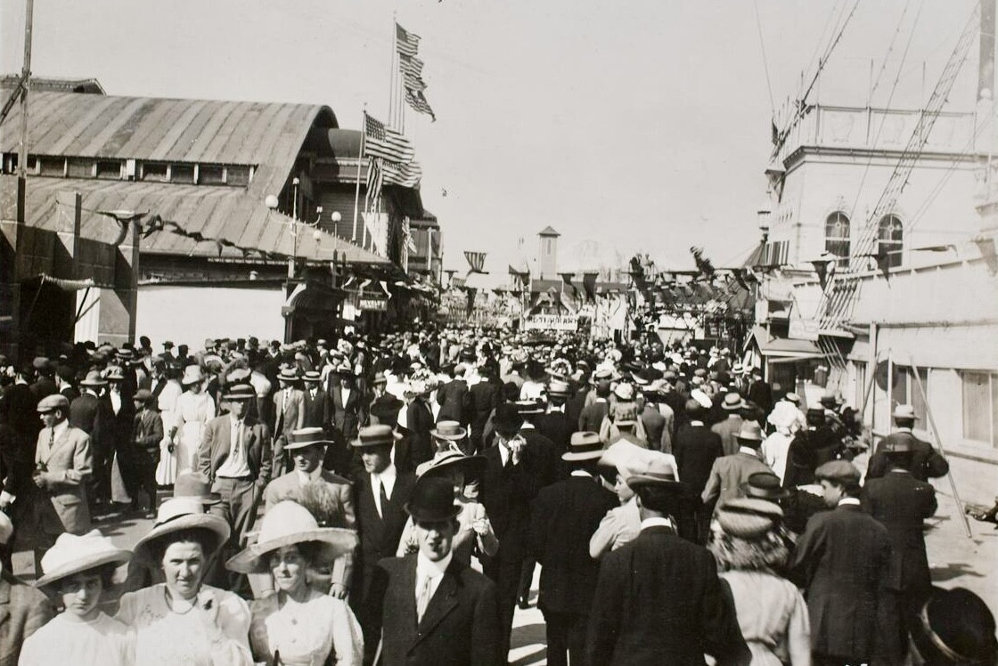 Black-and-white historic photograph of a large crowd on the Venice amusement pier around 1910, with people in hats walking between buildings and flags
