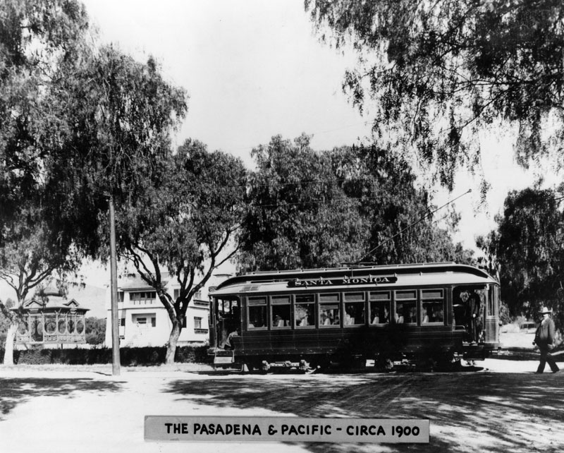 Black-and-white historic photograph of a Pasadena and Pacific streetcar around 1900, with passengers, trees and early homes nearby.