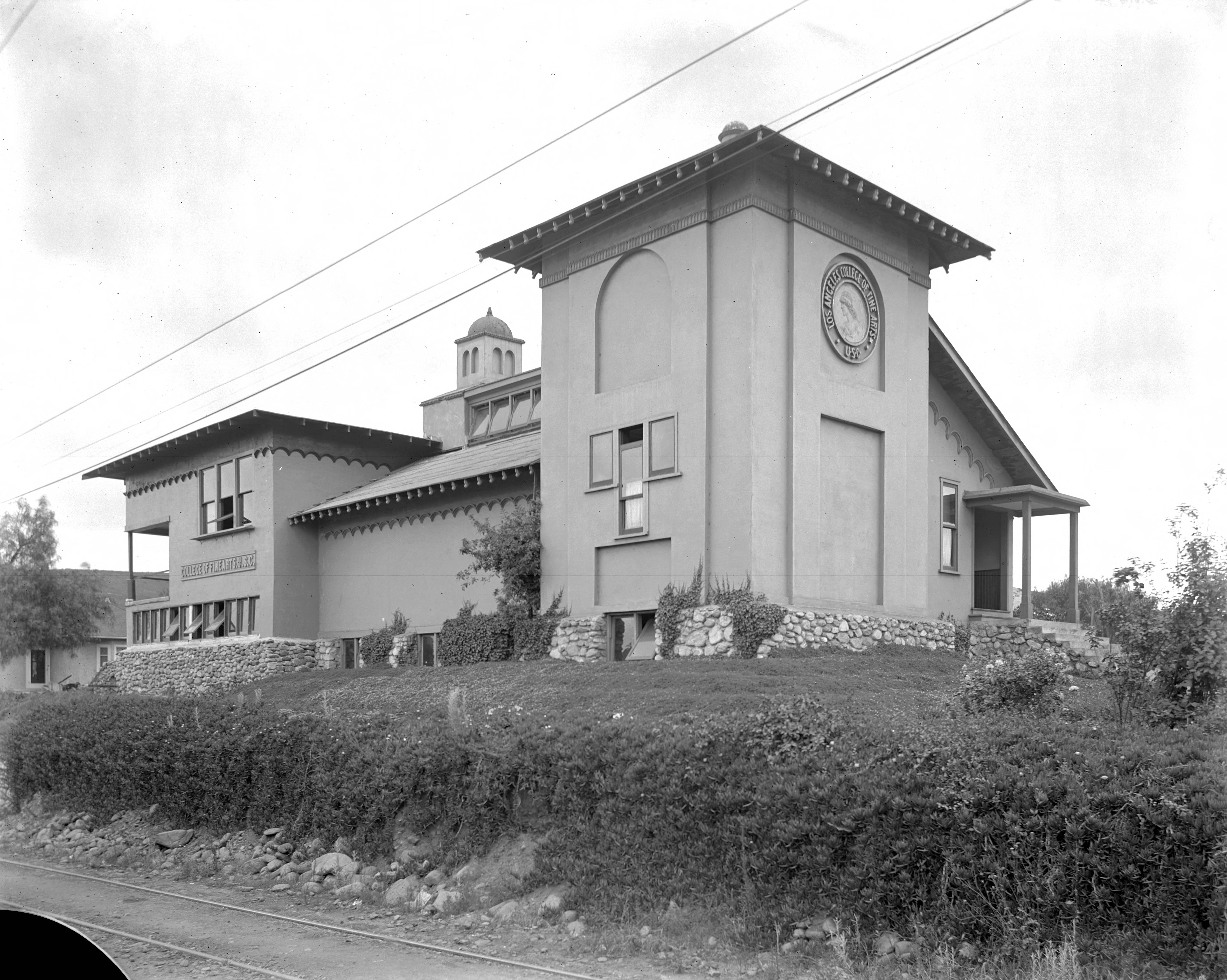  Black-and-white historic photograph of the Professor Judson College of Fine Arts building in Garvanza, with arched windows, a tower, stone base and hillside landscaping.