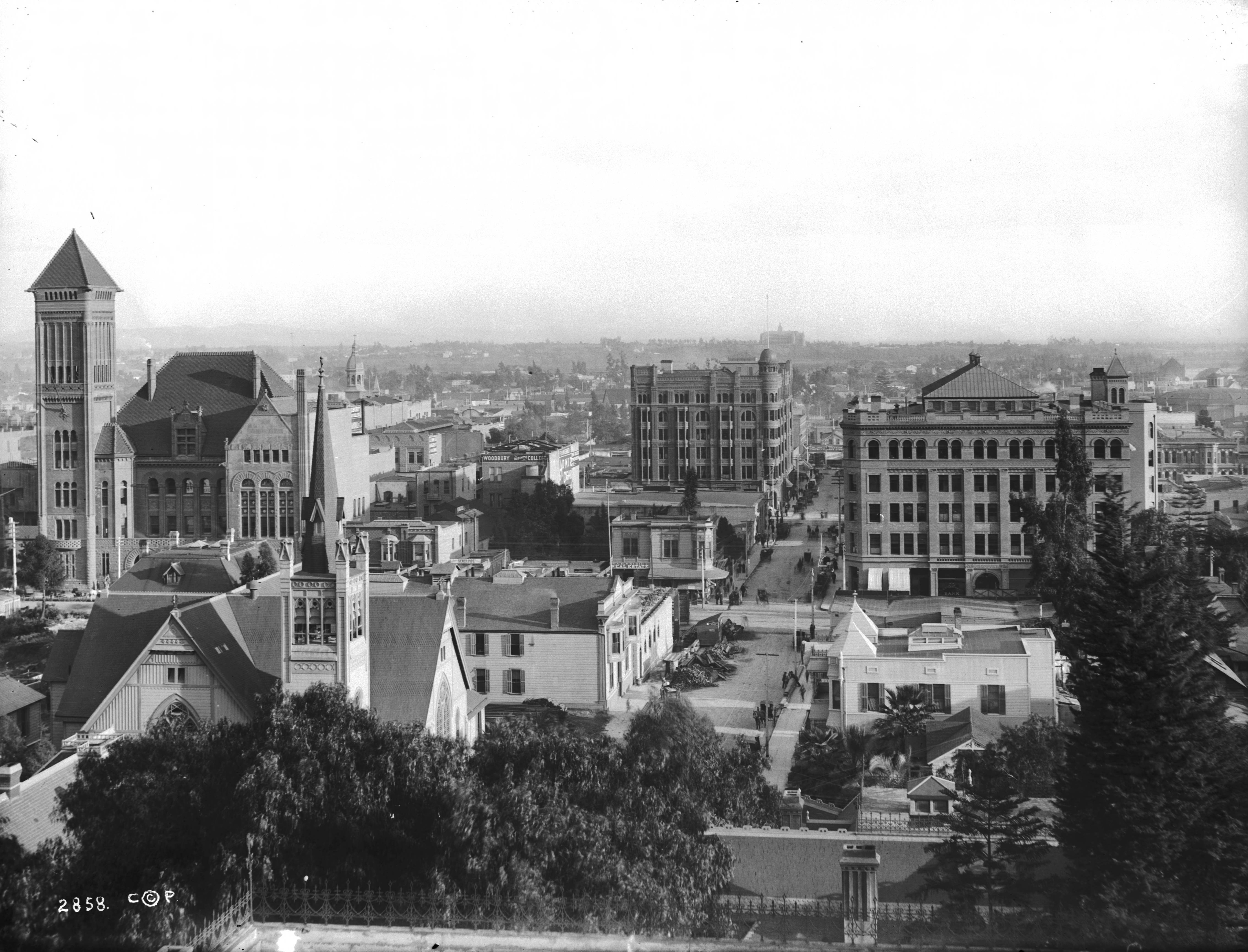 Broadway and Third Street in Downtown Los Angeles, showing the city's early commercial core. Black-and-white historic photograph of Broadway and Third Street in Downtown Los Angeles with early commercial buildings, churches, and streets visible from above.