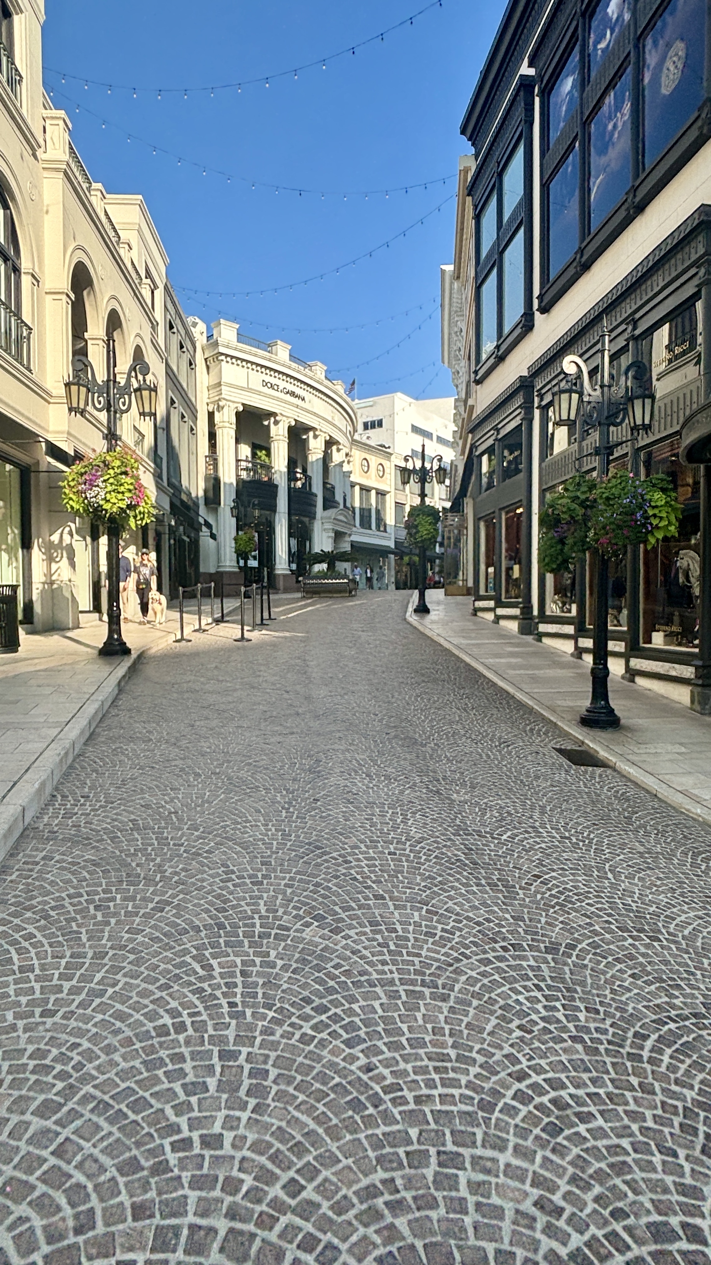 Via Rodeo in Beverly Hills with cobblestone paving, luxury storefronts, hanging lights and blue sky.