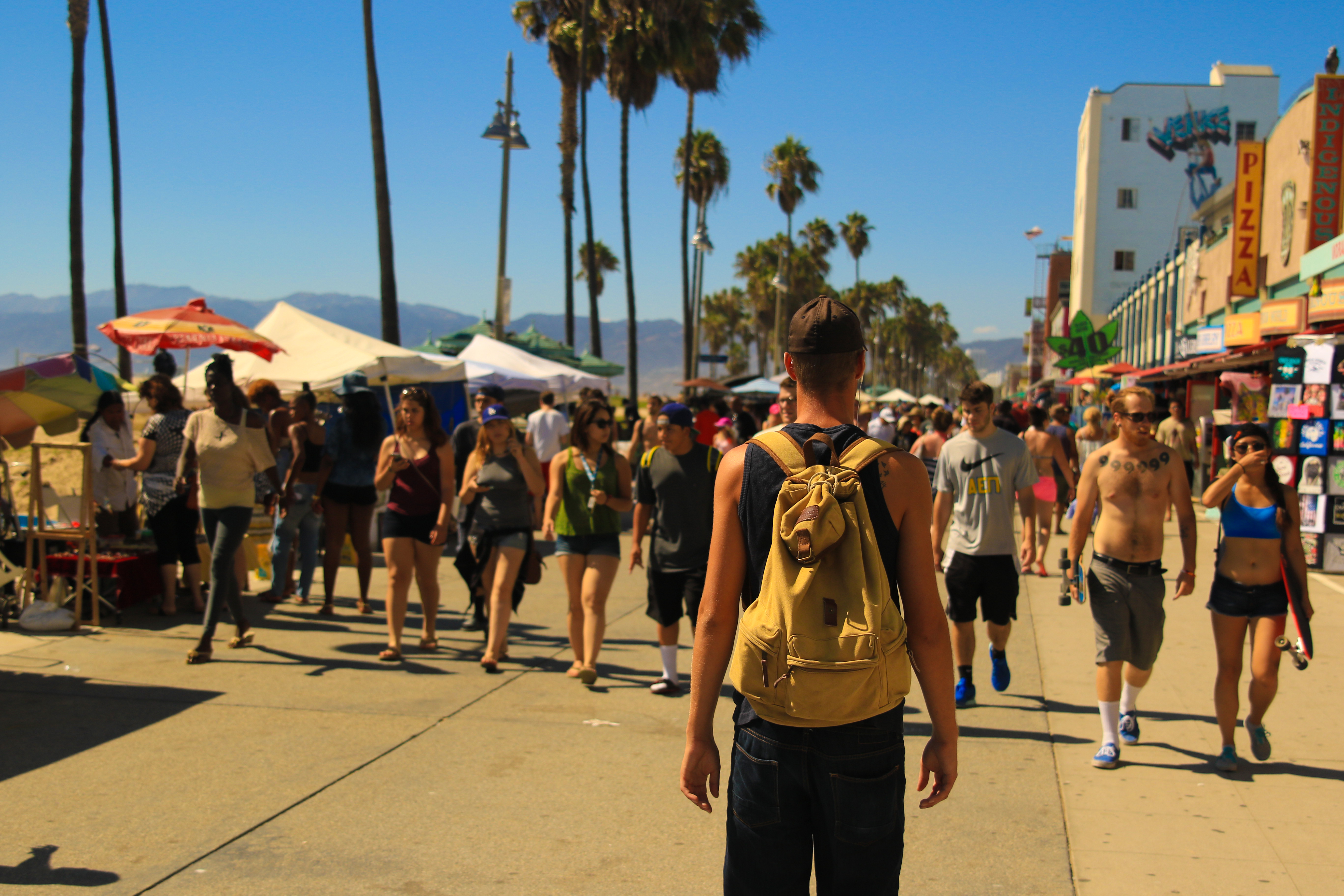 Crowded Venice Beach walkway with pedestrians, vendor tents, palm trees, beach shops, and mountains in the distance.