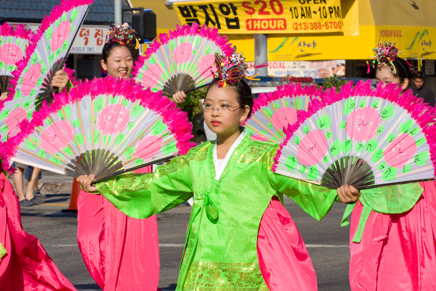 Korean cultural performers in bright traditional clothing holding large fans during a parade in Koreatown.
