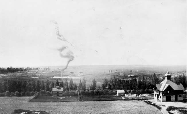 Early Inglewood, when the area was still transitioning from ranch land into a growing Southern California town. Black-and-white historic photograph of early Inglewood with open fields, scattered buildings, trees, and smoke rising in the distance.
