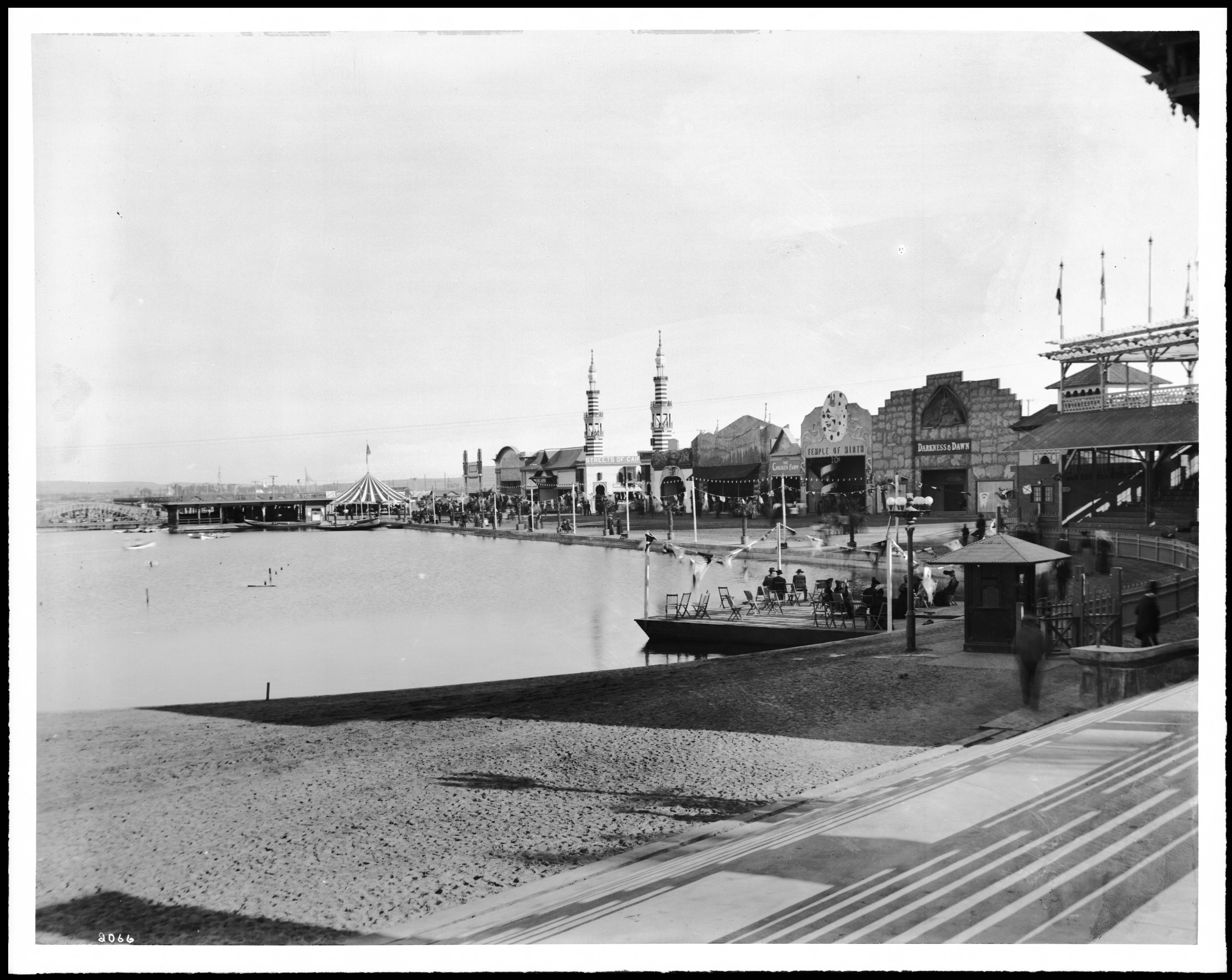 Black-and-white historic photograph of the Venice lagoon and beachfront businesses around 1910, with amusement buildings, water, steps, and people near the shoreline.