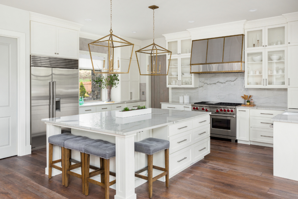 all white chef's kitchen with gold pendant lighting over large marble island and metal detailed range hood