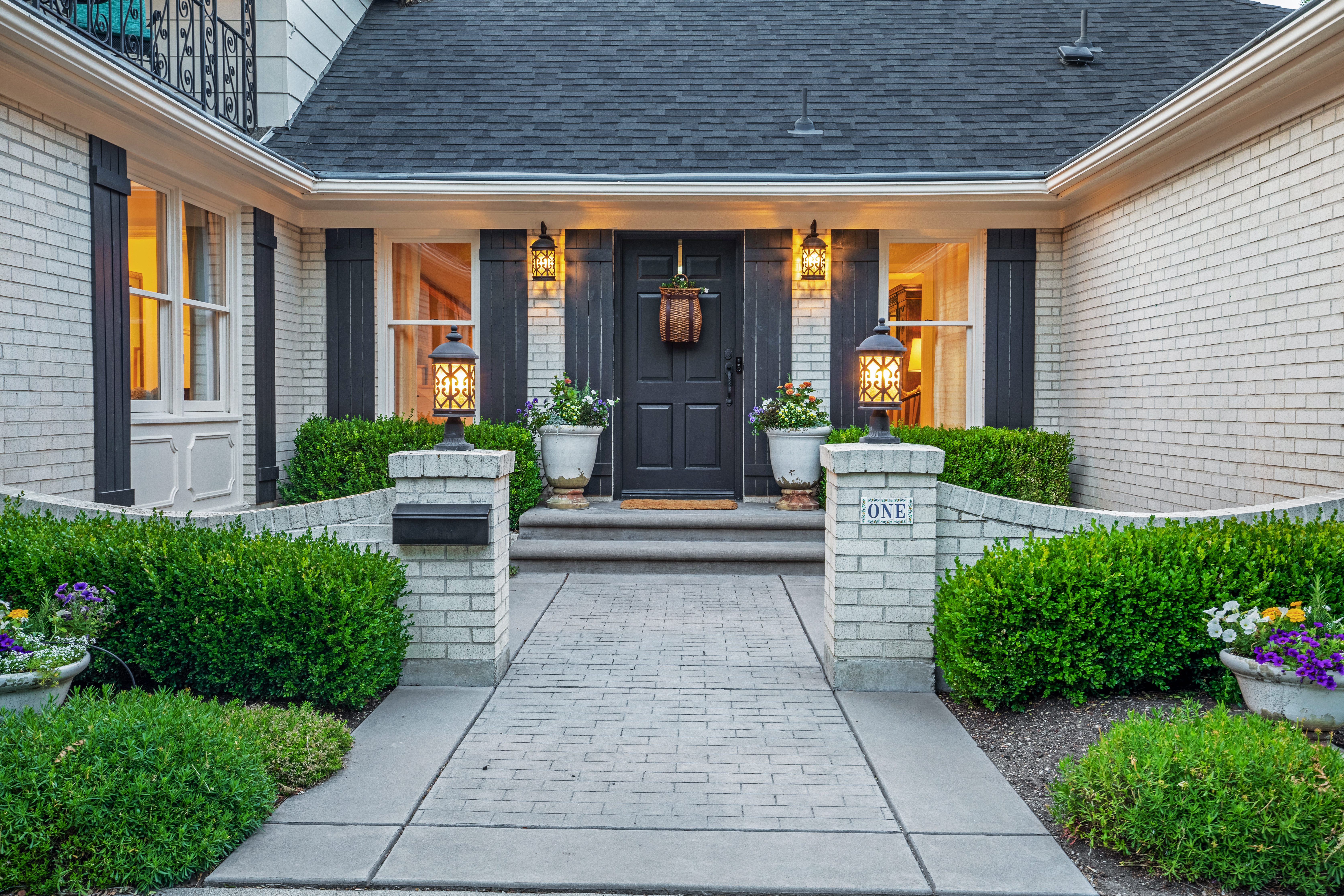 Inviting home entrance at dusk with warm porch lights, manicured bushes, and welcoming potted plants on either side of a dark wood door.