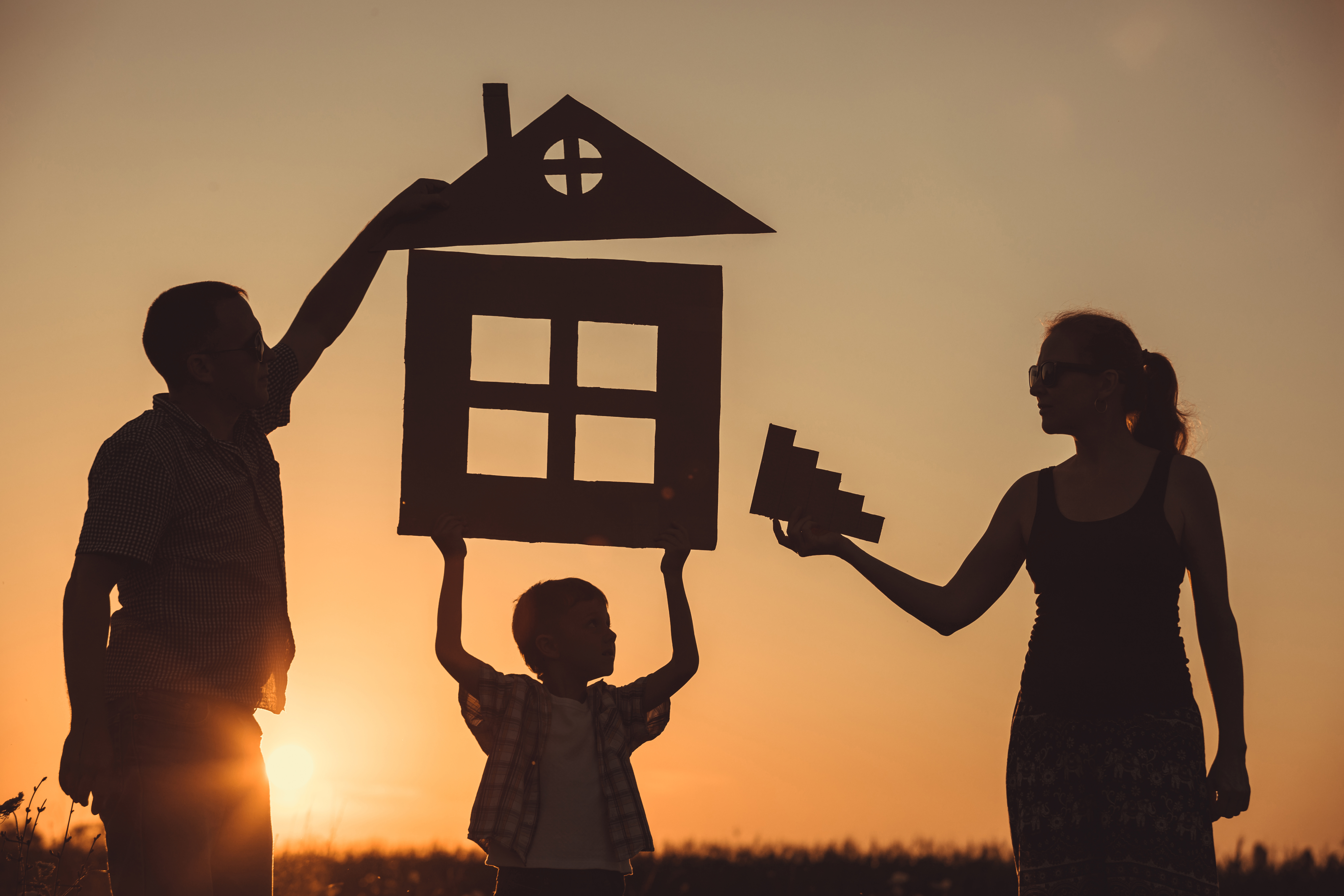 Silhouette of a family holding up cardboard cutouts of a house and stairs against a sunset, symbolizing the shared dream of homeownership.