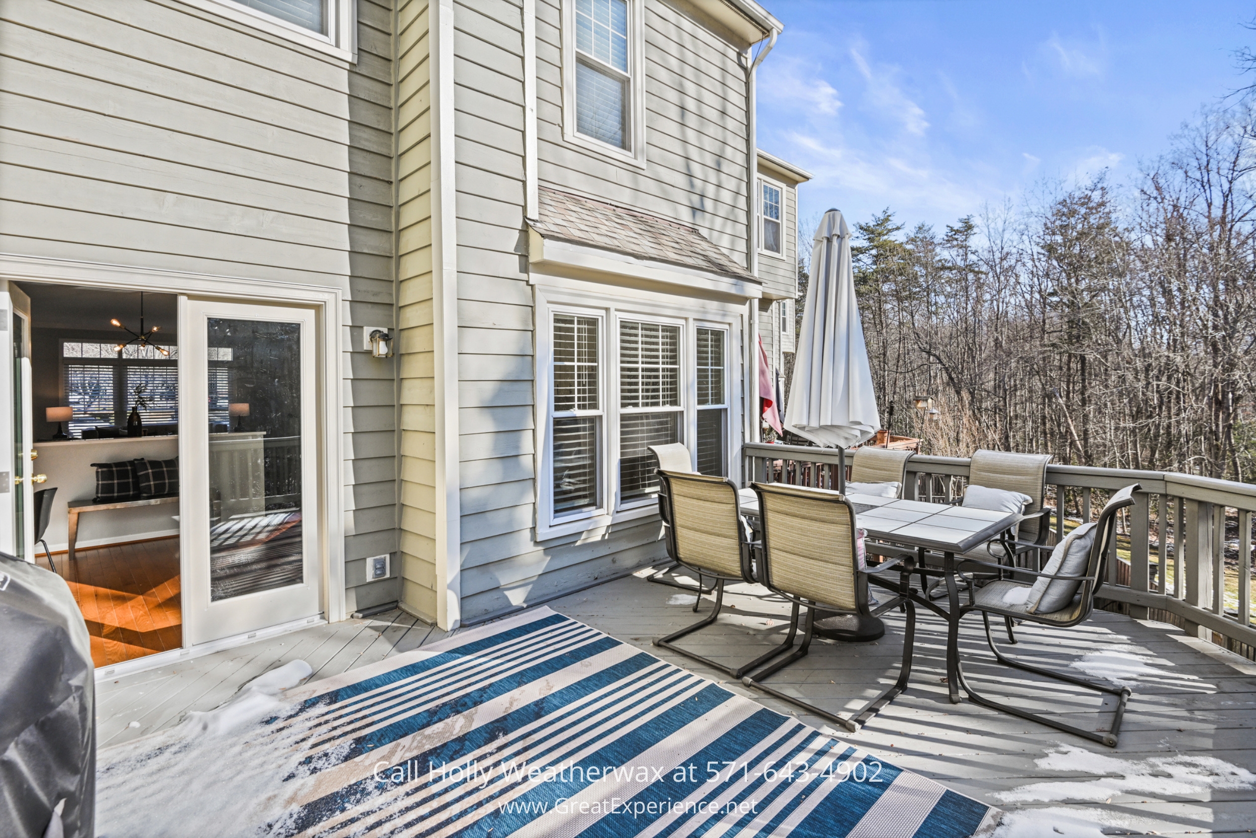 An outdoor deck with a dining table, chairs, and a blue-striped rug, providing a relaxing space with views of nearby trees and a clear blue sky.