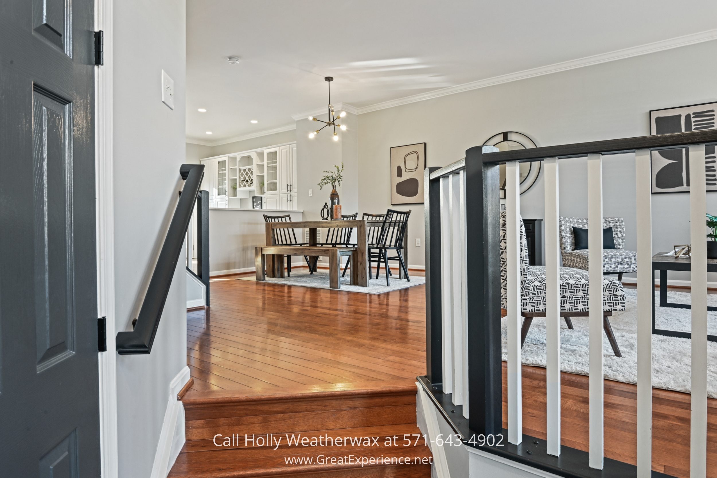 A bright entryway with warm hardwood flooring leading up to an open dining and living area, featuring a sleek black staircase railing.