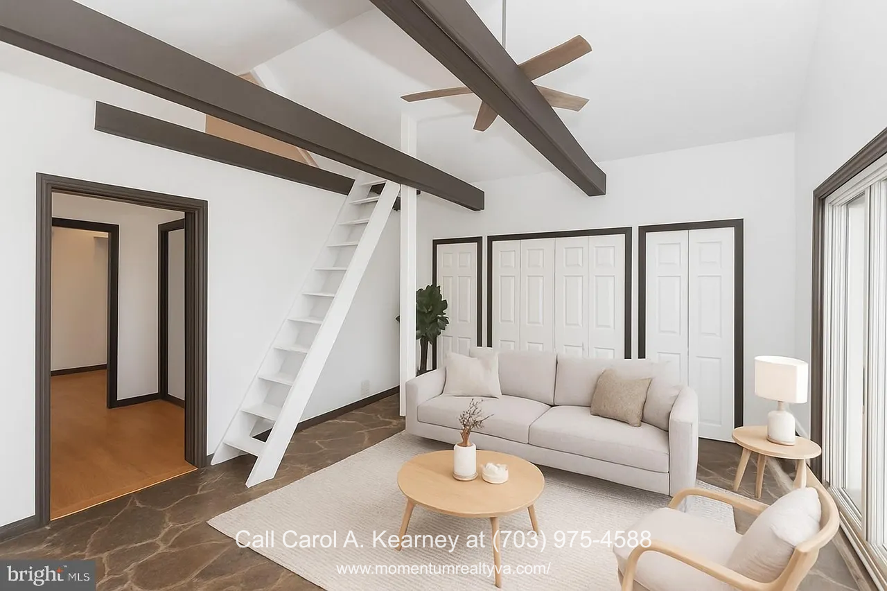 Bright living room inside the 420 White Oak Ln, Bluemont, VA home for sale, featuring vaulted ceilings with dark exposed beams, a white ladder leading to the loft storage area, stone flooring, and modern virtual staging with a beige sofa and round wooden coffee table.