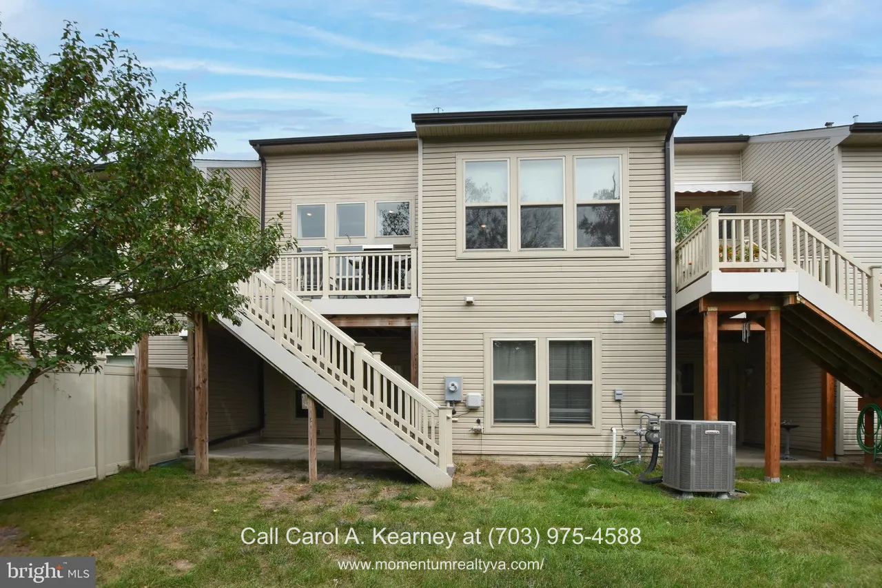 Rear exterior view of the townhouse at 19873 Silvery Blue Ter in Ashburn, VA, showing a main-level deck with stairs, walk-out lower level, and windows overlooking a private wooded setting in the Lexington 7 55+ community.