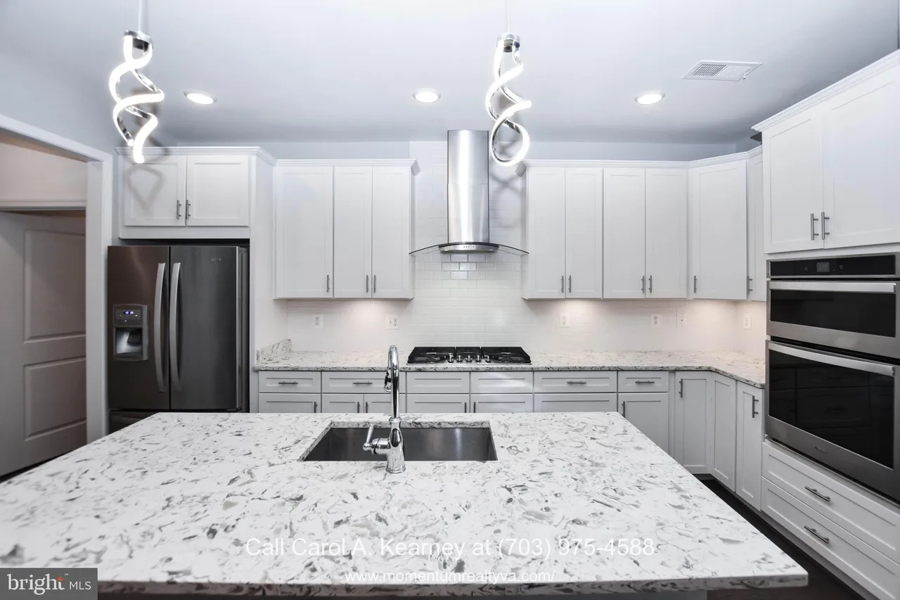 Gourmet kitchen at 19873 Silvery Blue Ter in Ashburn, VA, featuring white cabinetry, quartz countertops, a large center island with sink, stainless steel appliances, double wall ovens, and modern pendant lighting in the Lexington 7 55+ community.