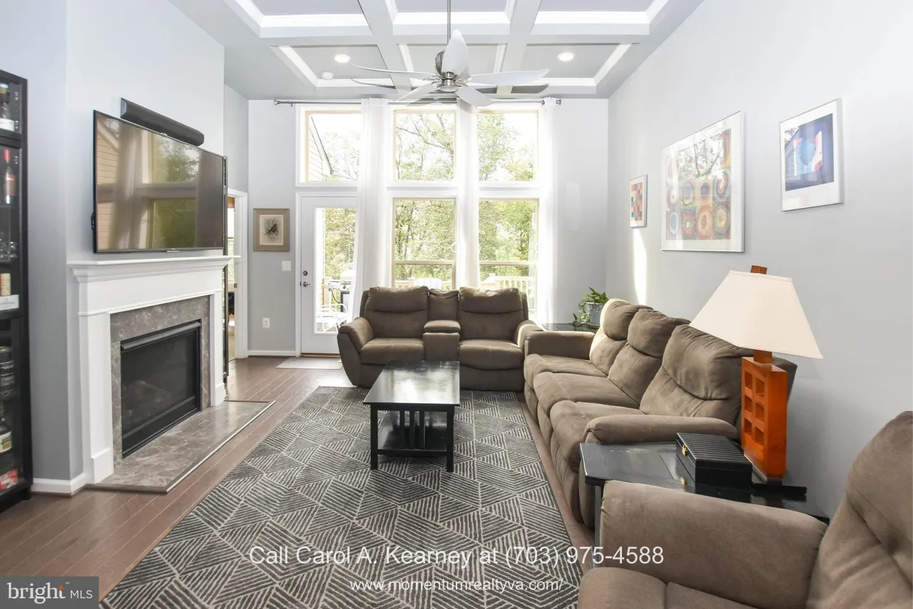 Bright open-concept living room at 19873 Silvery Blue Ter in Ashburn, VA, featuring a coffered ceiling with ceiling fan, large windows overlooking wooded views, gas fireplace, hardwood floors, and an inviting seating area in the Lexington 7 55+ community.
