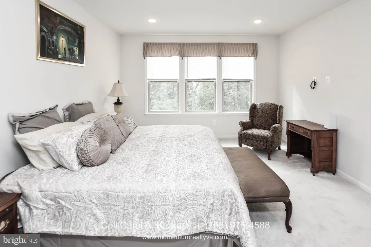 Main-level primary bedroom at 19873 Silvery Blue Ter in Ashburn, VA, featuring soft neutral tones, plush carpeting, large windows with wooded views, and a spacious layout designed for comfortable one-level living in the Lexington 7 55+ community.