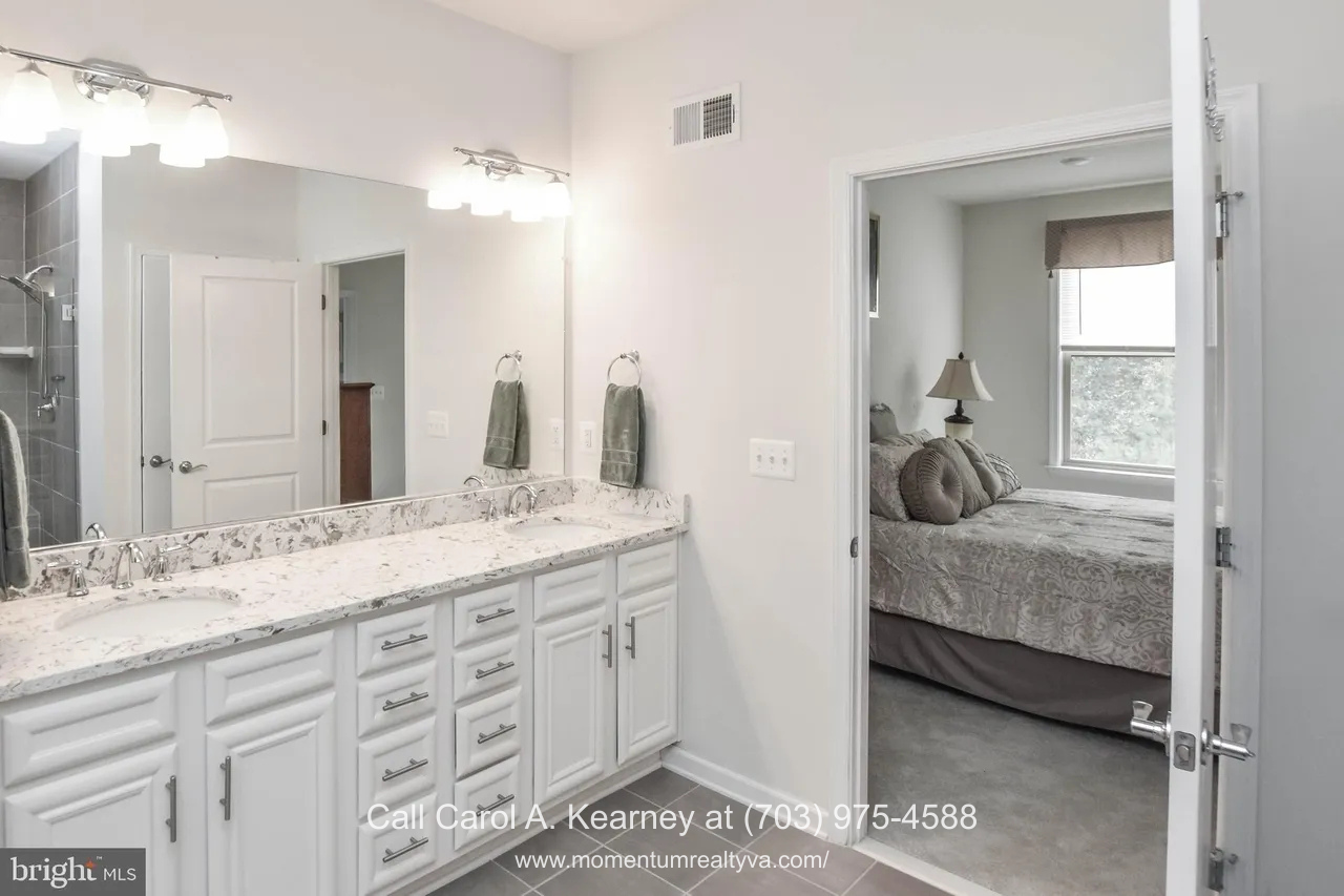 Main-level primary bathroom at 19873 Silvery Blue Ter in Ashburn, VA, featuring a double vanity with quartz countertops, white cabinetry, large mirror, modern lighting, and direct access to the primary bedroom in the Lexington 7 55+ community.