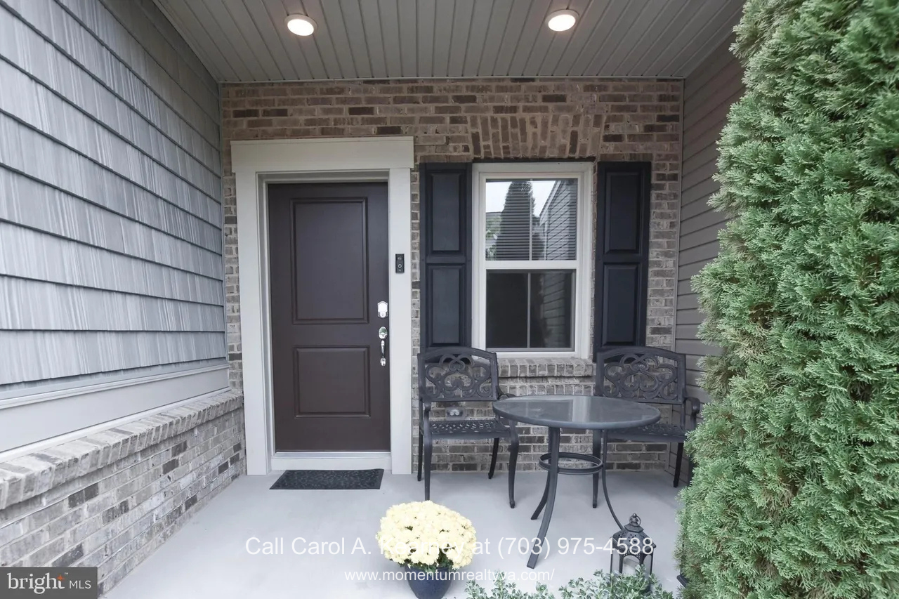 Covered front porch of a Lexington 7 townhouse at 19873 Silvery Blue Ter in Ashburn, VA, featuring a brick façade, dark front door, recessed lighting, and a small patio seating area overlooking landscaped greenery in a 55+ community.